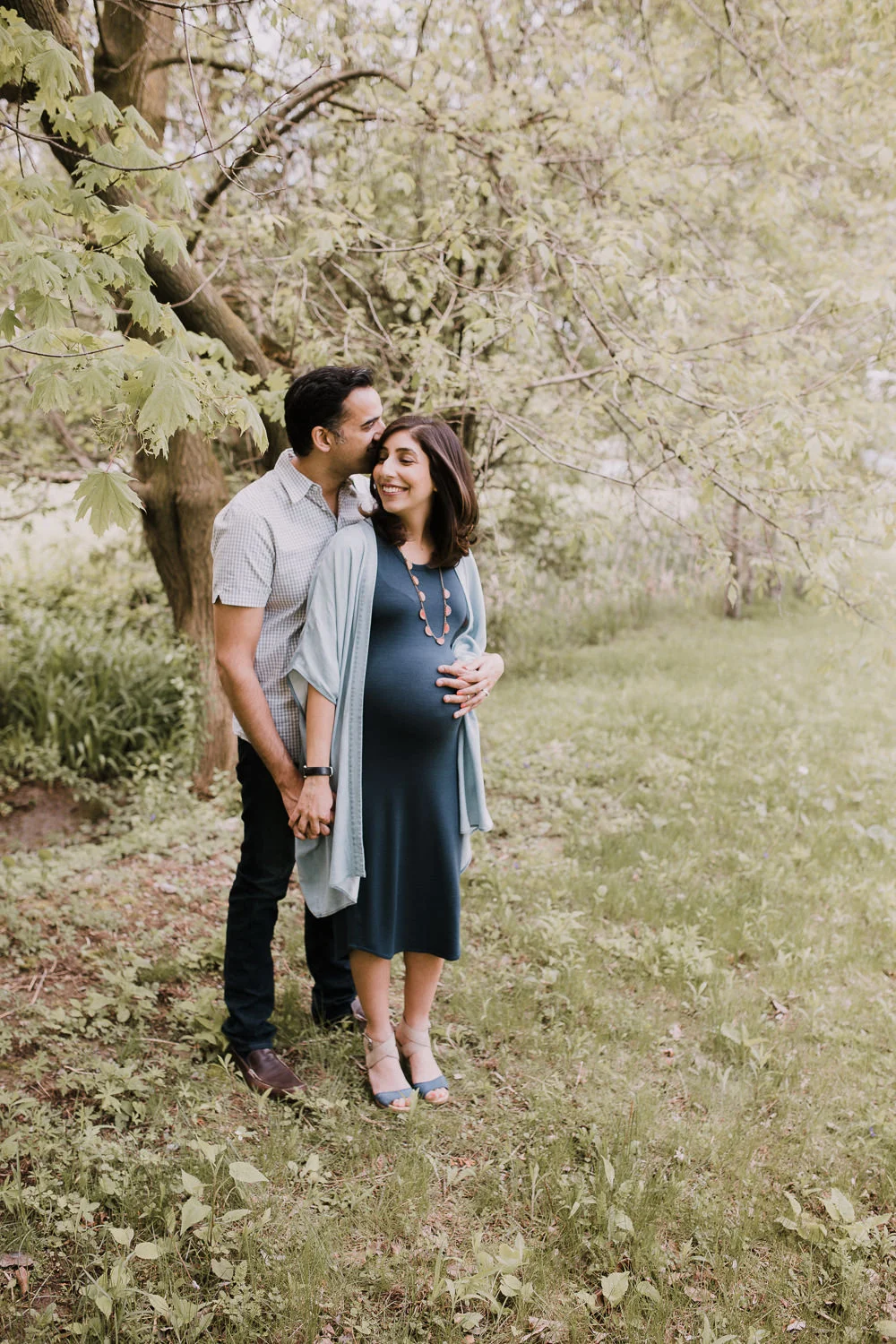 husband standing behind pregnant wife, holding her hand and baby bump and kissing her head - Markham Lifestyle Photography