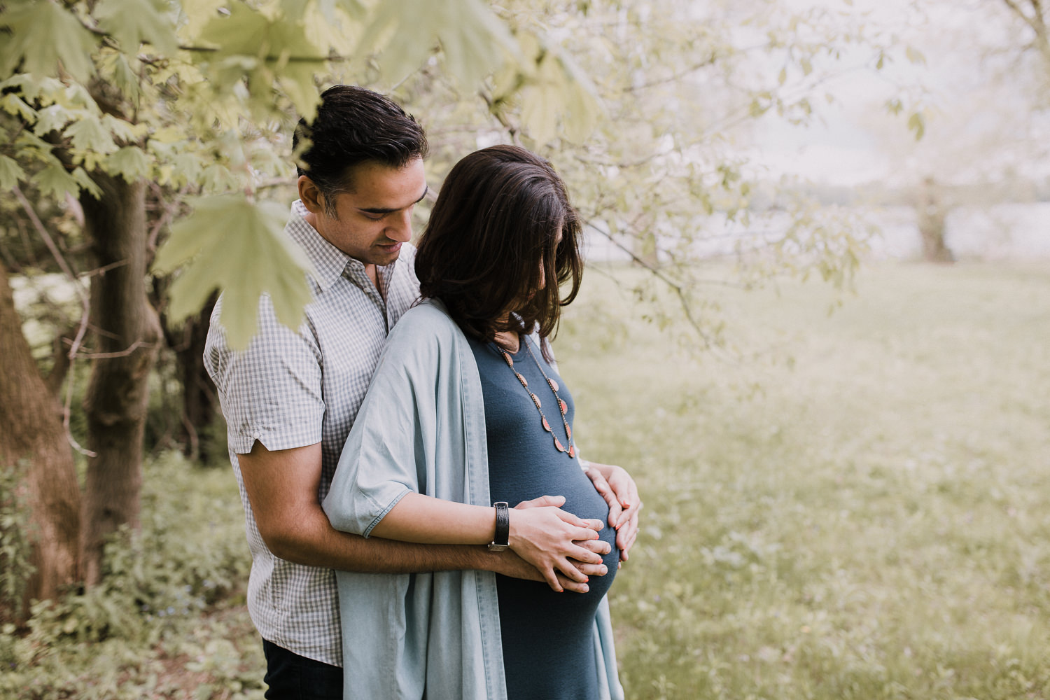 husband embracing pregnant wife from behind, parents-to-be looking down at baby bump - Stouffville In-Home Photography