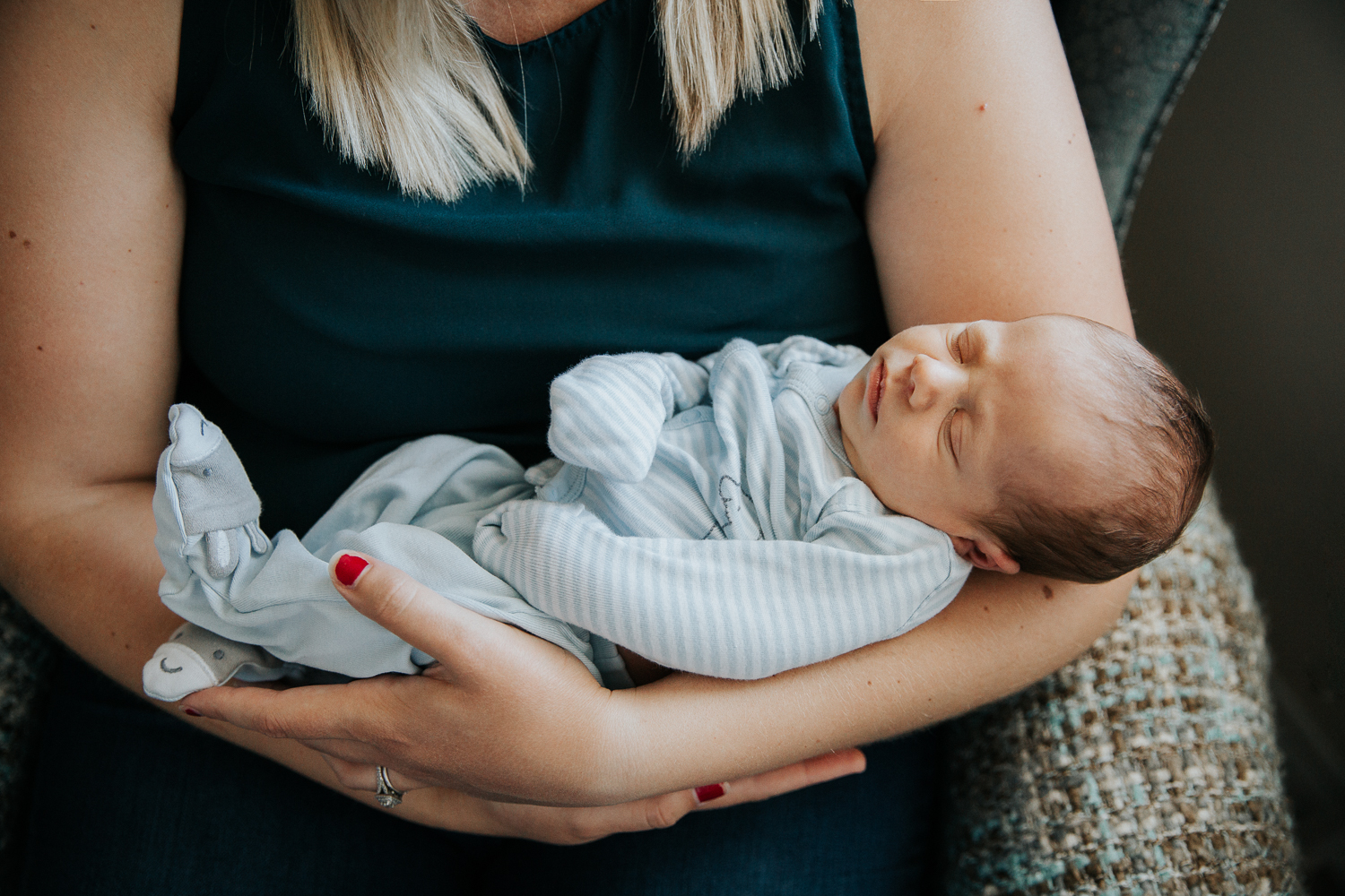 new mom sitting in nursery chair holding 2 week old baby boy in blue sleeper - Stouffville Lifestyle Photography