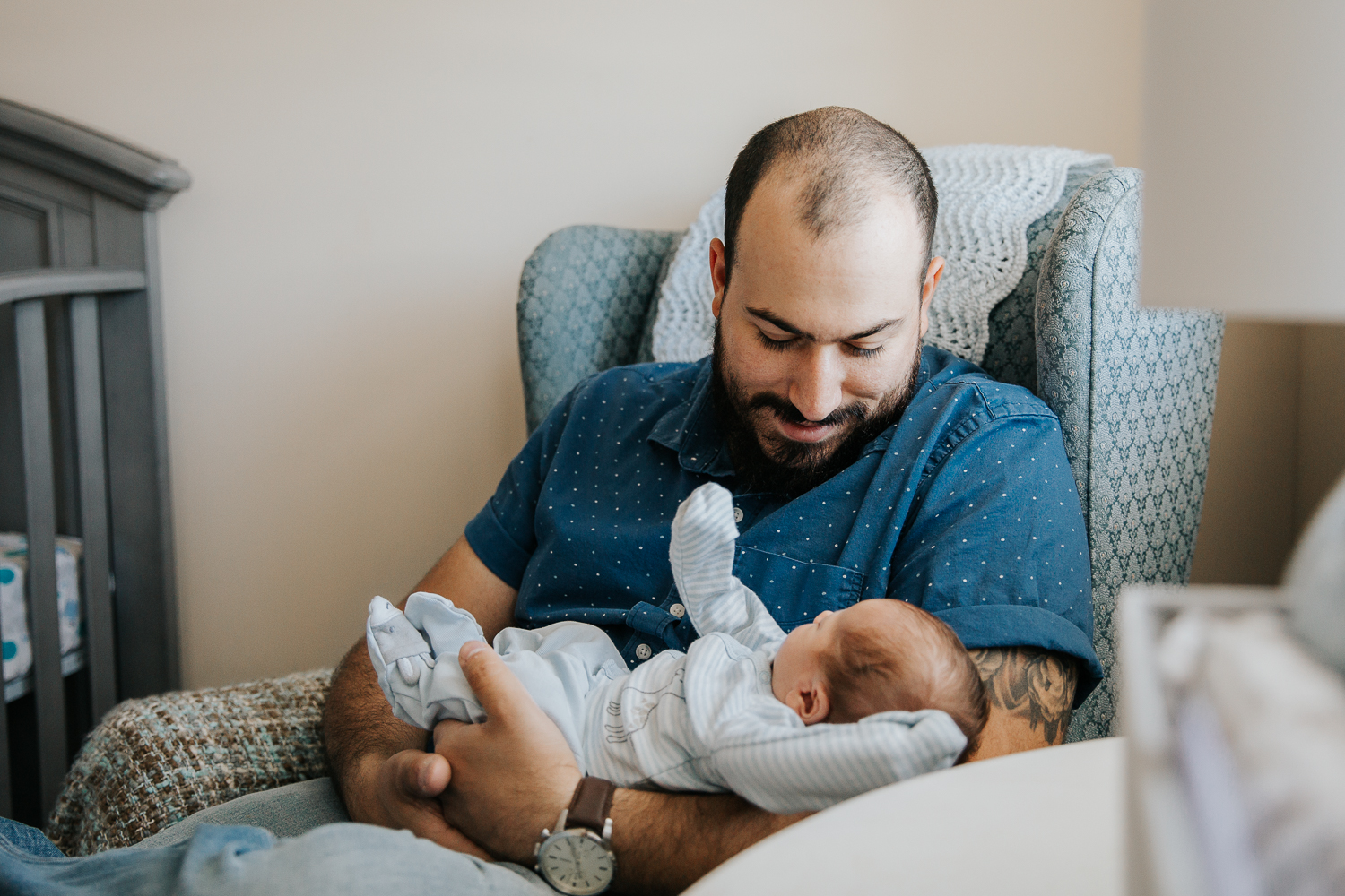 new dad sitting in nursery chair holding and looking at 2 week old baby boy in blue sleeper - Newmarket In-Home Photography