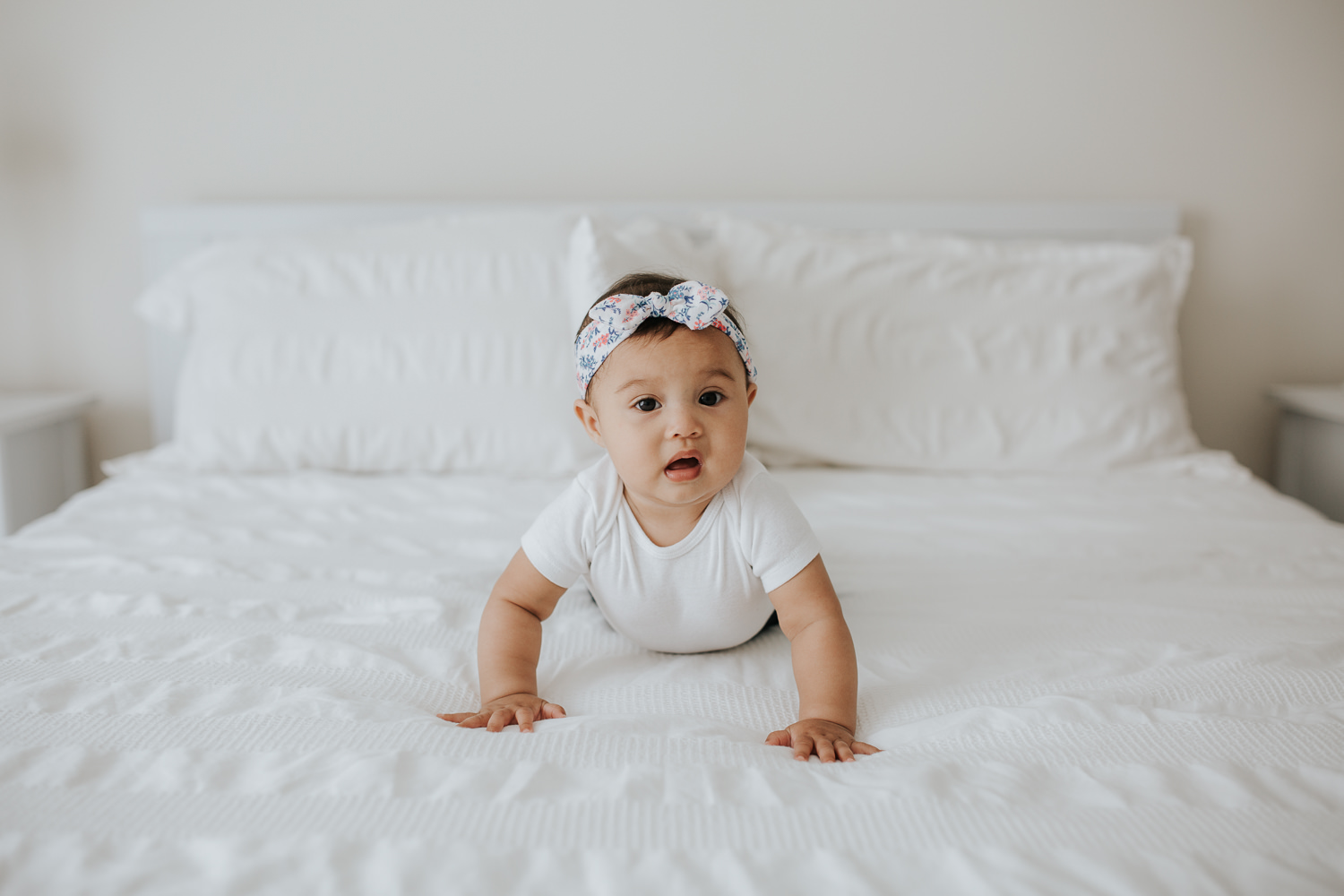 6 month old baby girl with floral headband on crawling on white bed, looking at camera -  Newmarket Lifestyle Photography