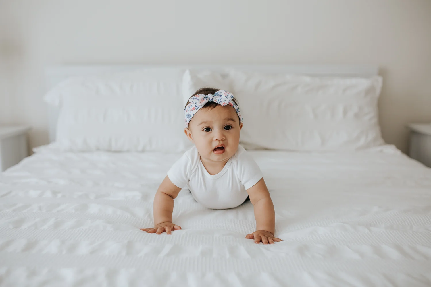 6 month old baby girl with floral headband on crawling on white bed, looking at camera - Newmarket Lifestyle Photography