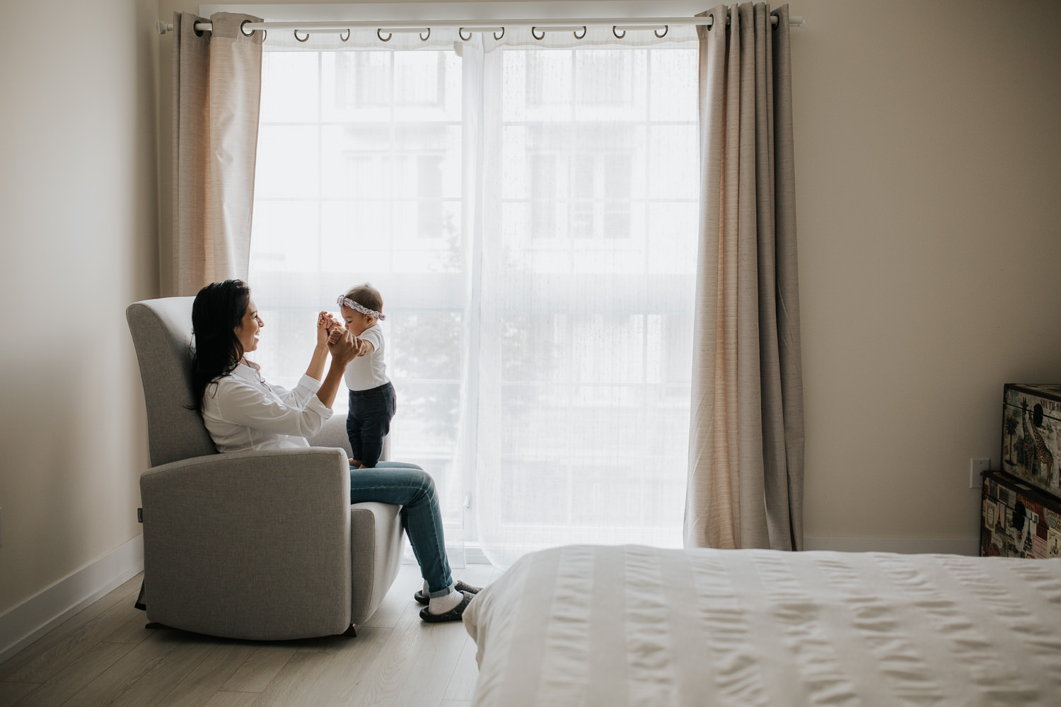 6 month old baby girl standing on mother's legs, mom sitting on chair holding daughter's hands - Markham In-Home Photography