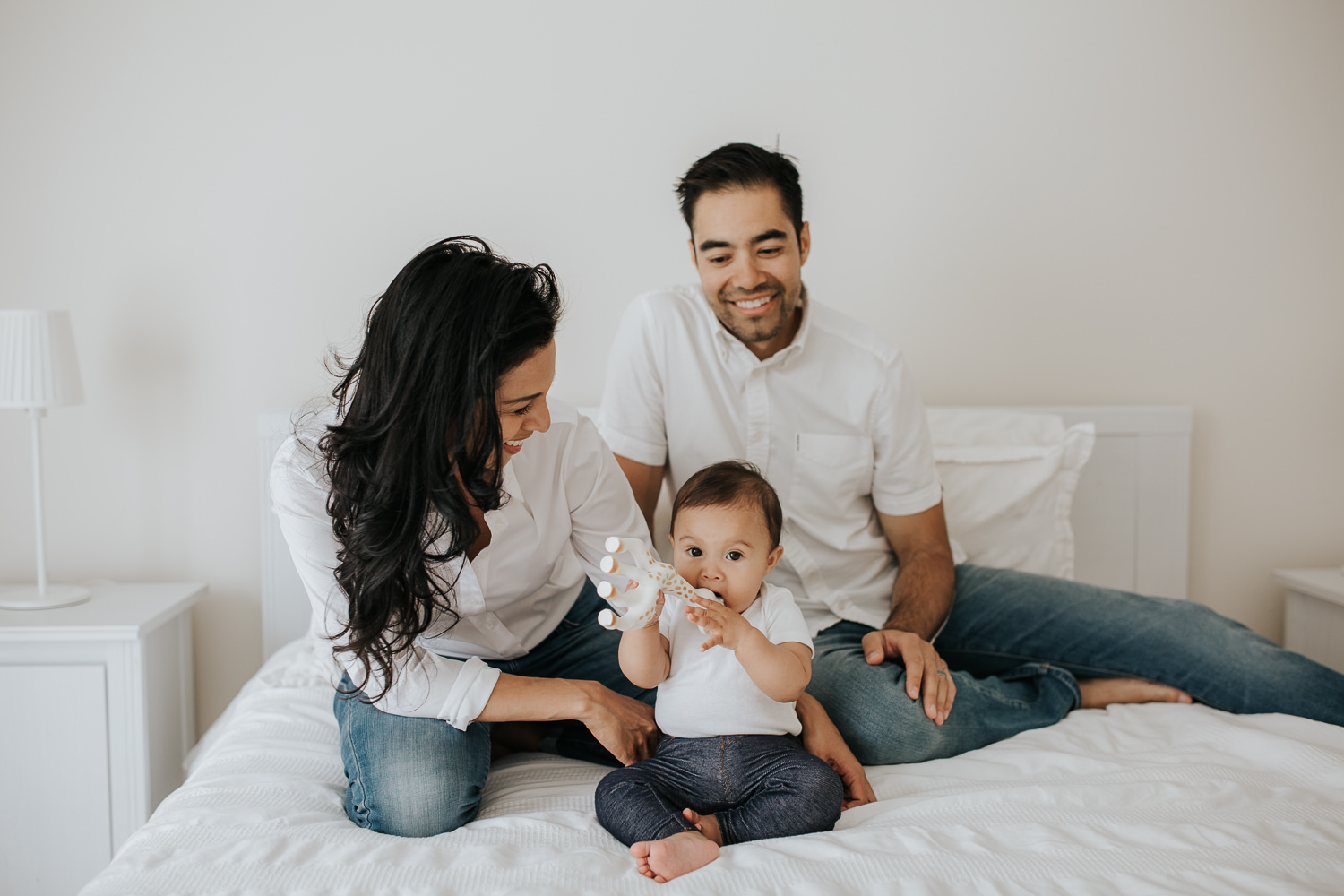 6 month old baby girl sitting on bed chewing Sophie the giraffe, while parents sit behind her - Newmarket Lifestyle Photos