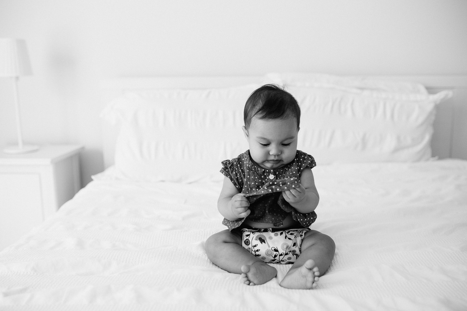 6 month old baby girl with dark hair sits on bed holding and looking at dress - Newmarket Lifestyle Photography