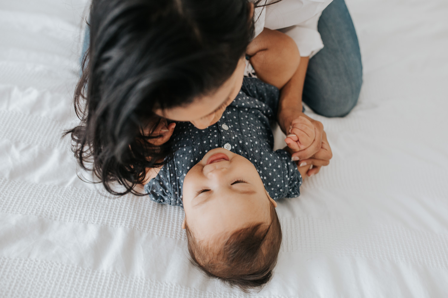 smiling 6 month old baby girl lying on bed, mom leaning over her playing- Barrie Lifestyle Photography