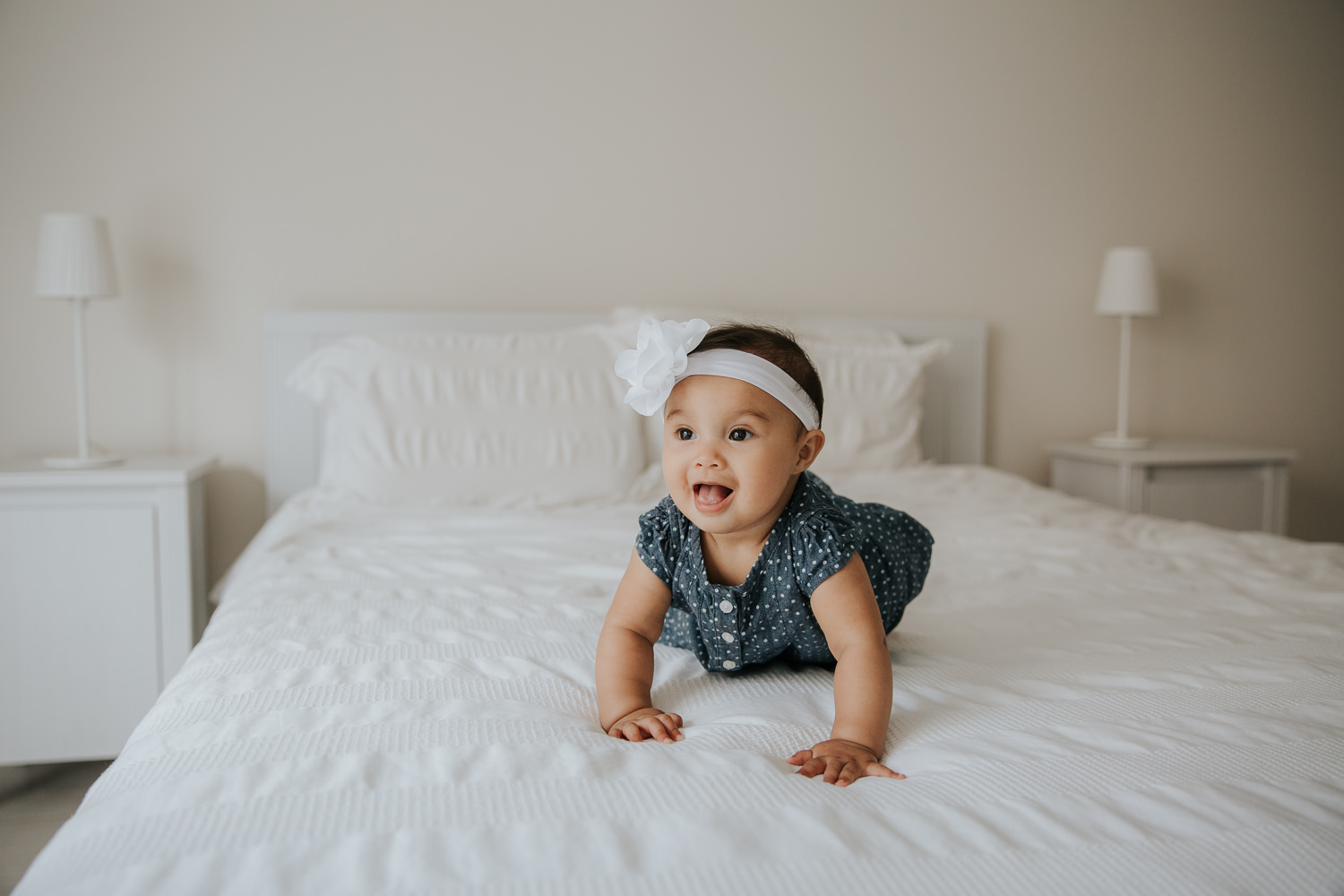 6 month old baby girl with dark hair and eyes in polka dot dress and headband crawling on bed, smiling -  Newmarket In-Home Photography