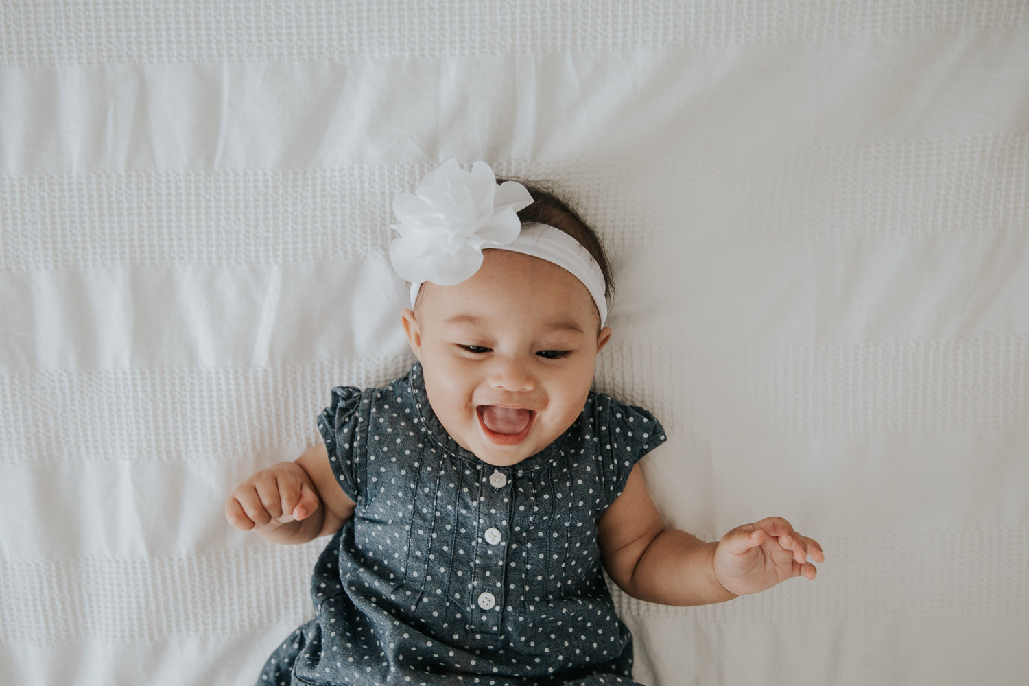 6 month old baby girl in blue and white polka dot dress lying on bed laughing - Markham In-Home Photos