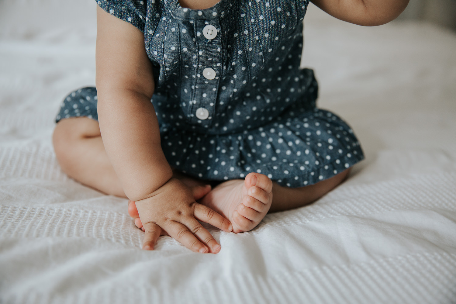 6 month old baby girl in blue and white polka dot dress sitting on bed, close up of hand and feet - Newmarket Lifestyle Photos
