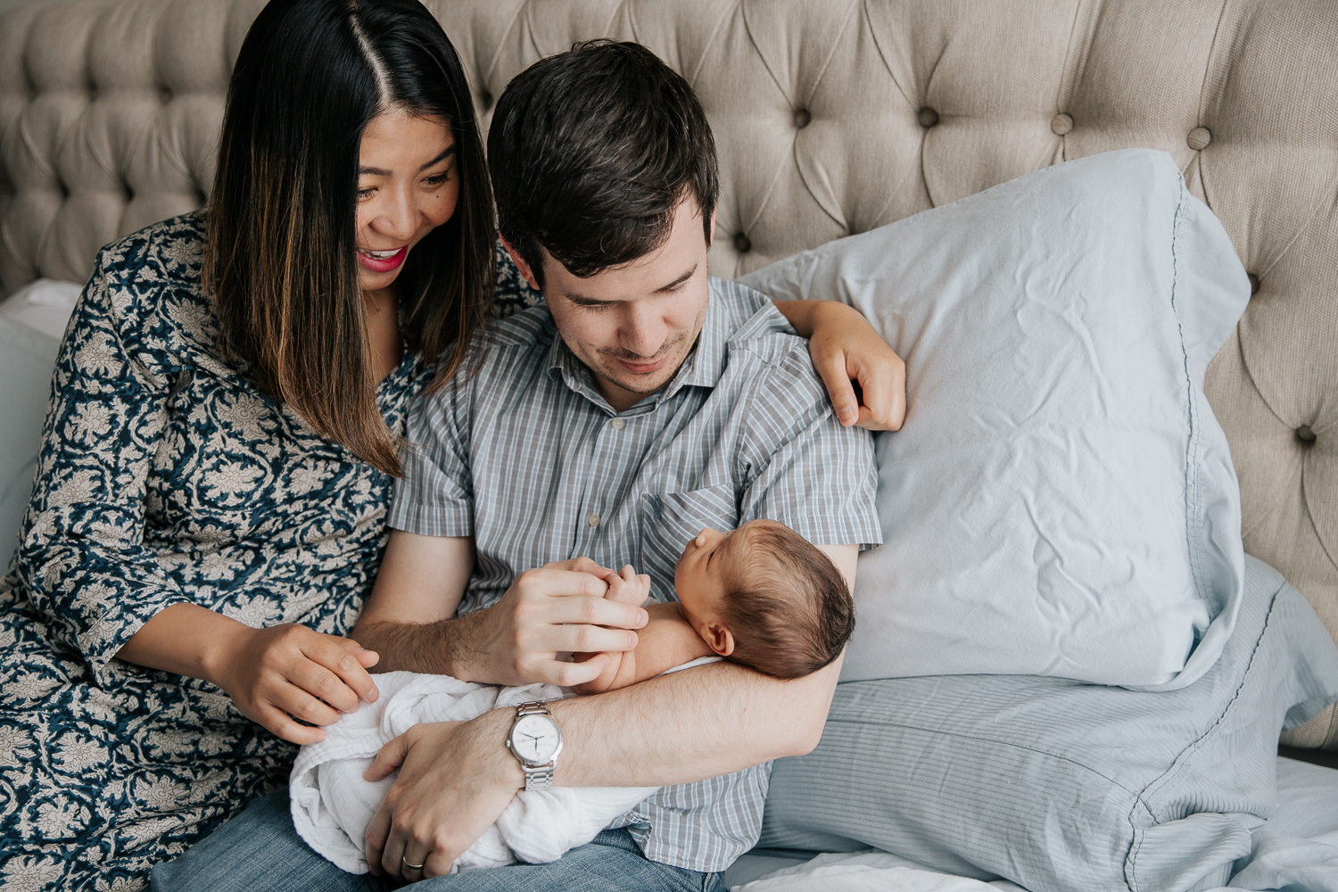 new parents sitting on master bed holding and smiling at 2 week old baby girl - Newmarket In-Home Photography