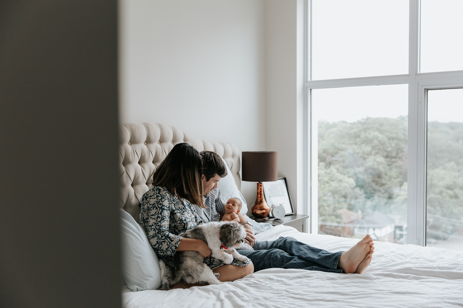 new parents sitting on master bed holding 2 week old baby girl and shitzu dog - Stouffville Lifestyle Photography