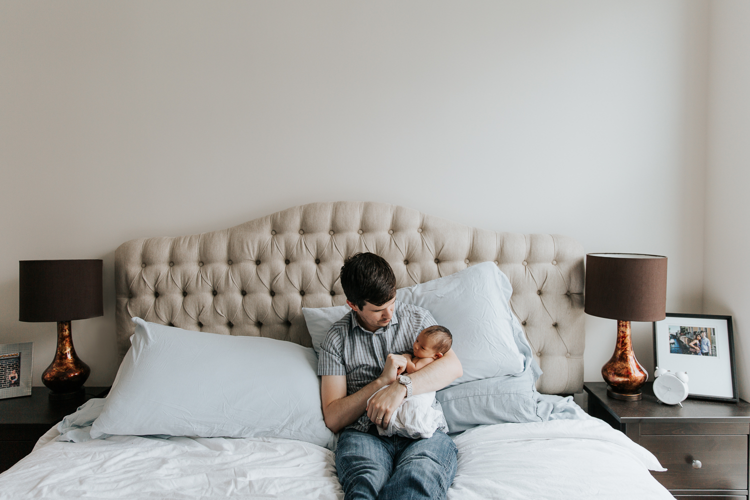 new father sitting on master bed holding swaddled 2 week old baby girl - Newmarket In-Home Photos