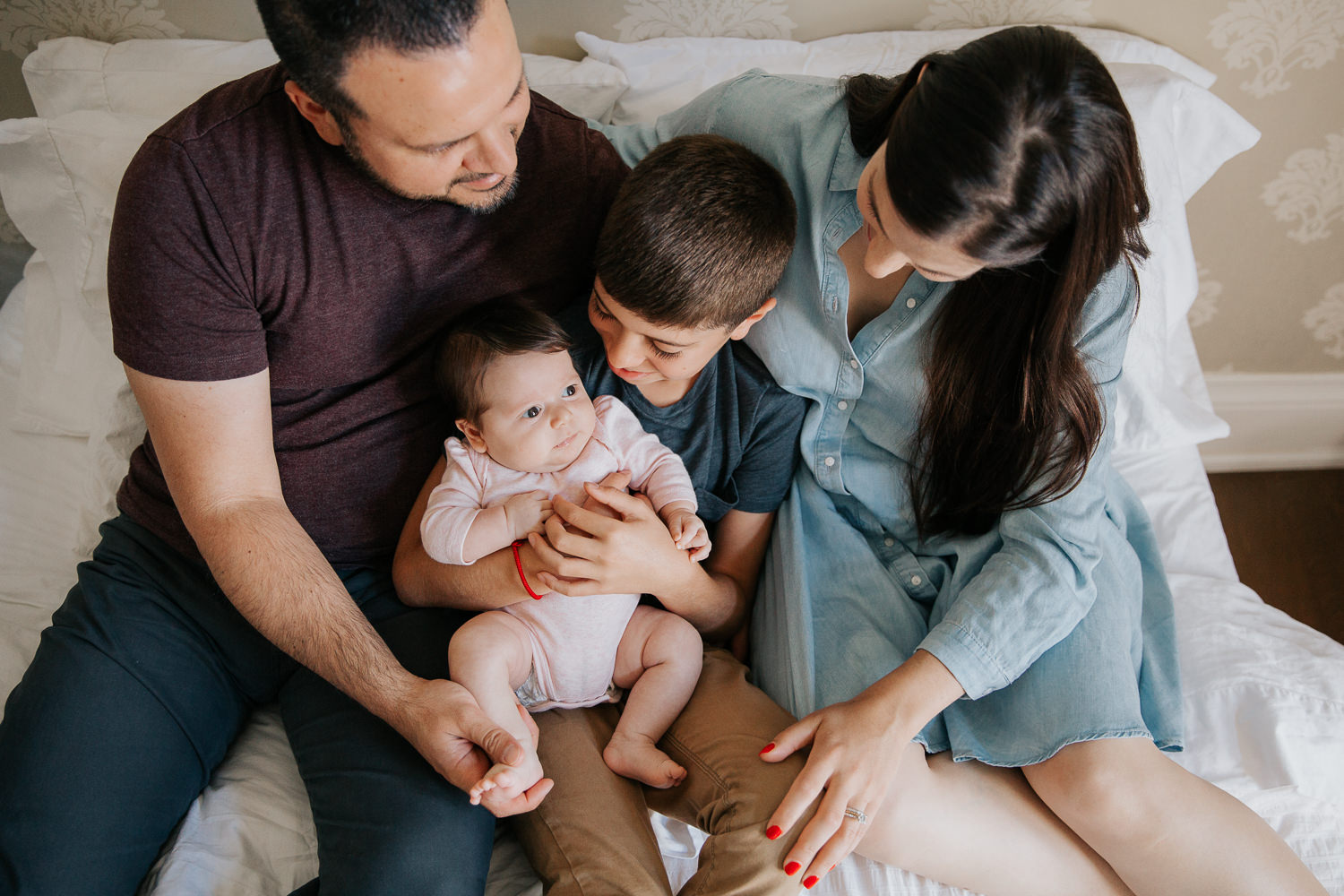 family of four sitting on bed, 9 year old boy holding 2 month old baby sister in pink onesie as mom and dad snuggled next to them - Newmarket Lifestyle Photography
