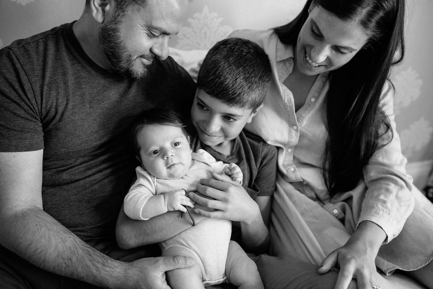 family of four sitting on bed, 9 year old boy holding 2 month old baby sister in onesie as mom and dad snuggled next to them - Stouffville Lifestyle Photography