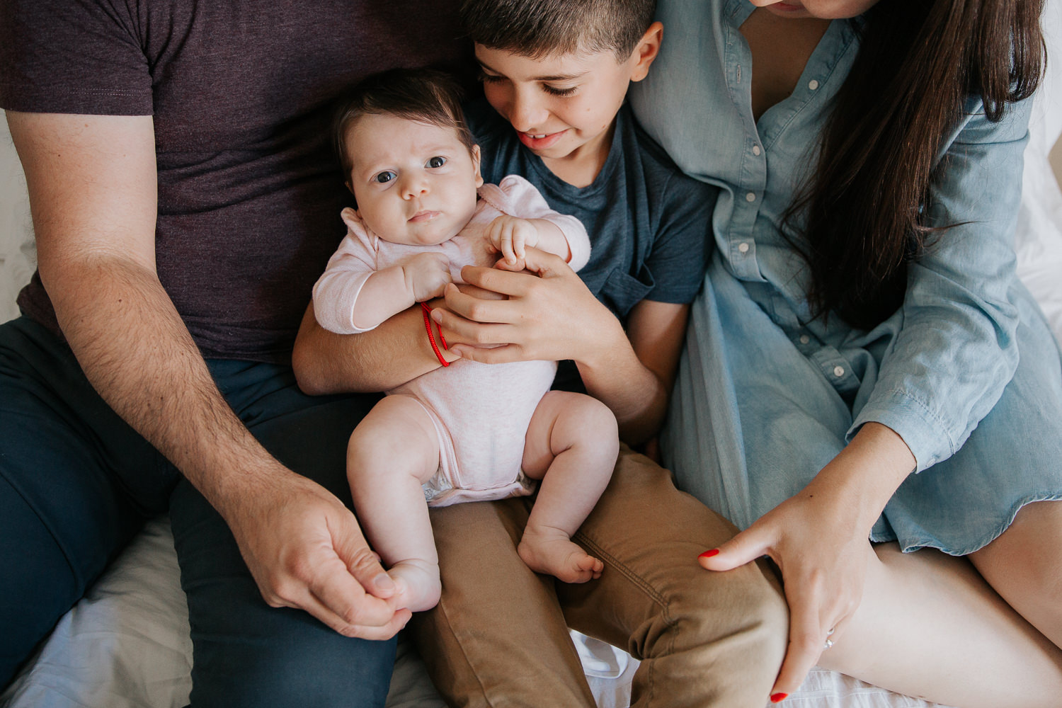 family of four sitting on bed, 9 year old boy holding 2 month old baby sister in pink onesie as mom and dad snuggled next to them - Markham Lifestyle Photography