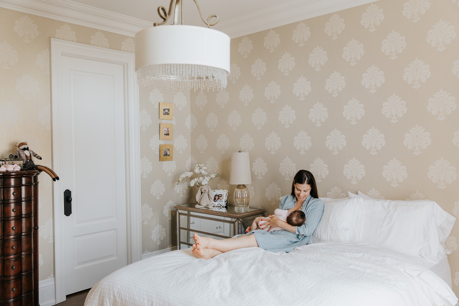 new mom with dark hair in blue chambray dress sitting on master bed, nursing and smiling at 2 month old baby girl in her arms - Stouffville In-Home Photography