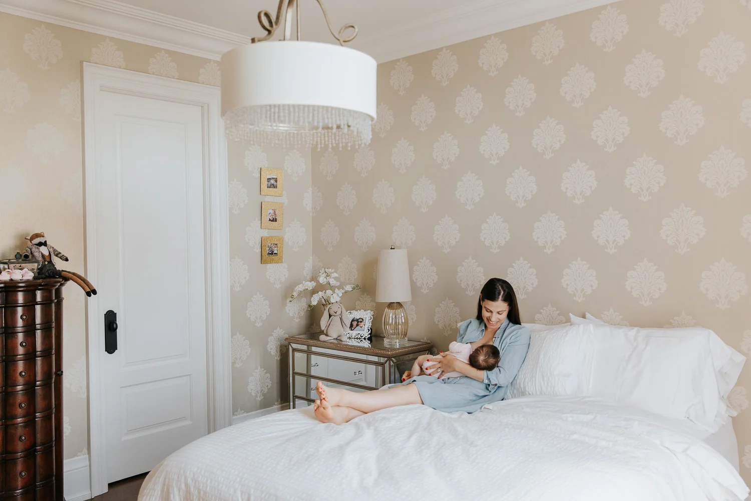 new mom with dark hair in blue chambray dress sitting on master bed, nursing and smiling at 2 month old baby girl in her arms - Stouffville In-Home Photography
