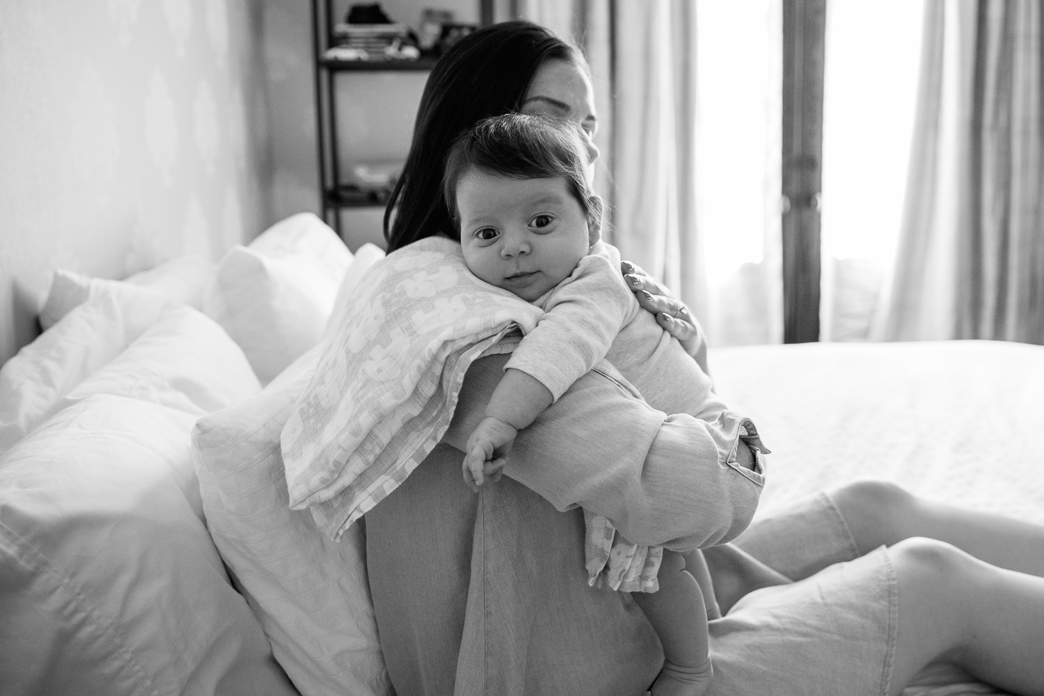 new mom with dark hair in blue chambray dress sitting on master bed, holding 2 month old daughter over her should, burping her, as baby girl looks at the camera - Markham In-Home Photography