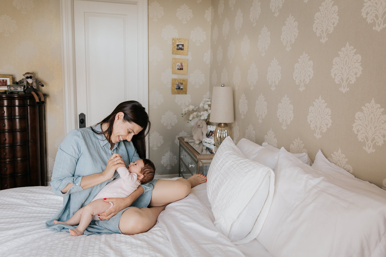 first time mother with long brunette hair in blue chambray dress sitting on master bed, smiling at 2 month old baby girl in her arms and holding her hand - Newmarket In-Home Photos