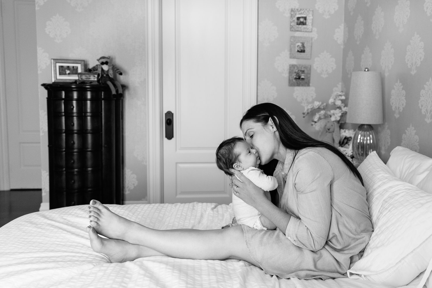 first time mother with long brunette hair in blue chambray dress sitting on master bed, kissing 2 month old baby girl on the cheek - Stouffville In-Home Photos