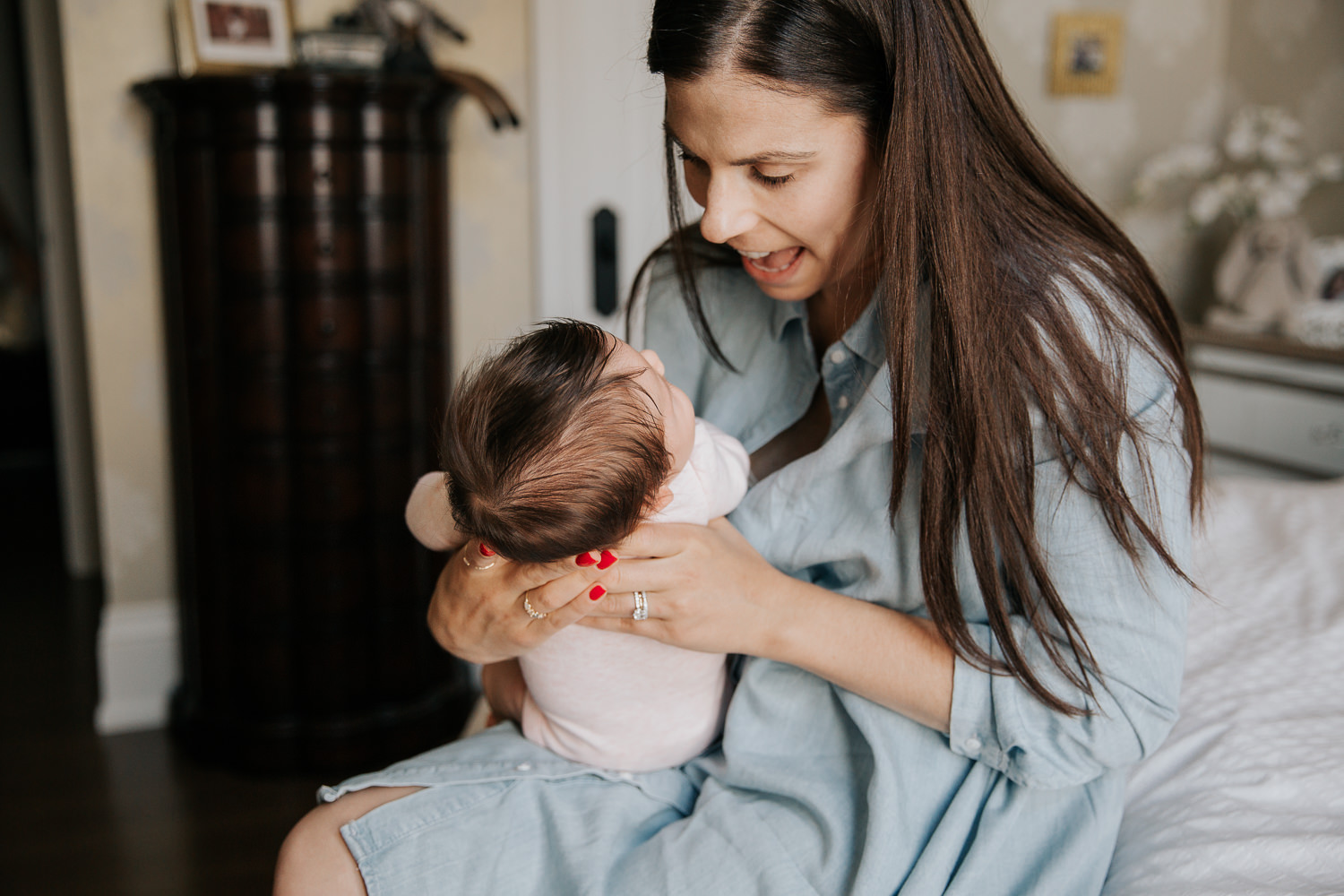 first time mother with long brunette hair in blue chambray dress sitting on master bed, smiling at 2 month old baby girl in pink onesie her arms - Markham In-Home Photos
