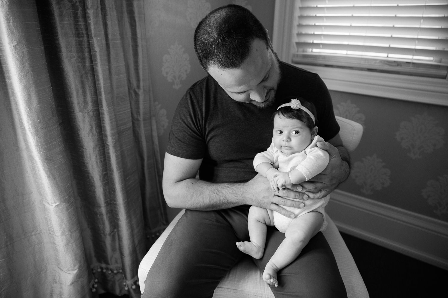 new dad in jeans and t shirt sitting in chair holding and smiling at 2 month old baby girl in onesie who is sitting on his knee looking at the camera -  GTA In-Home Photos