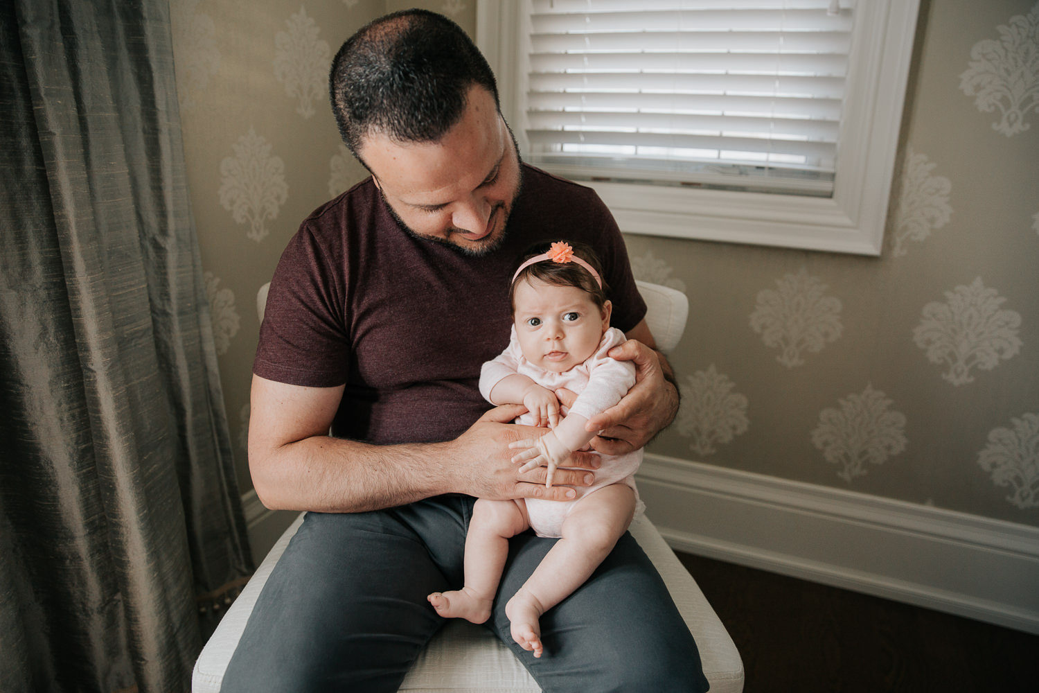 new dad in jeans and t shirt sitting in chair holding and smiling at 2 month old baby girl in pink onesie who is sitting on his knee looking at the camera - Barrie In-Home Photos