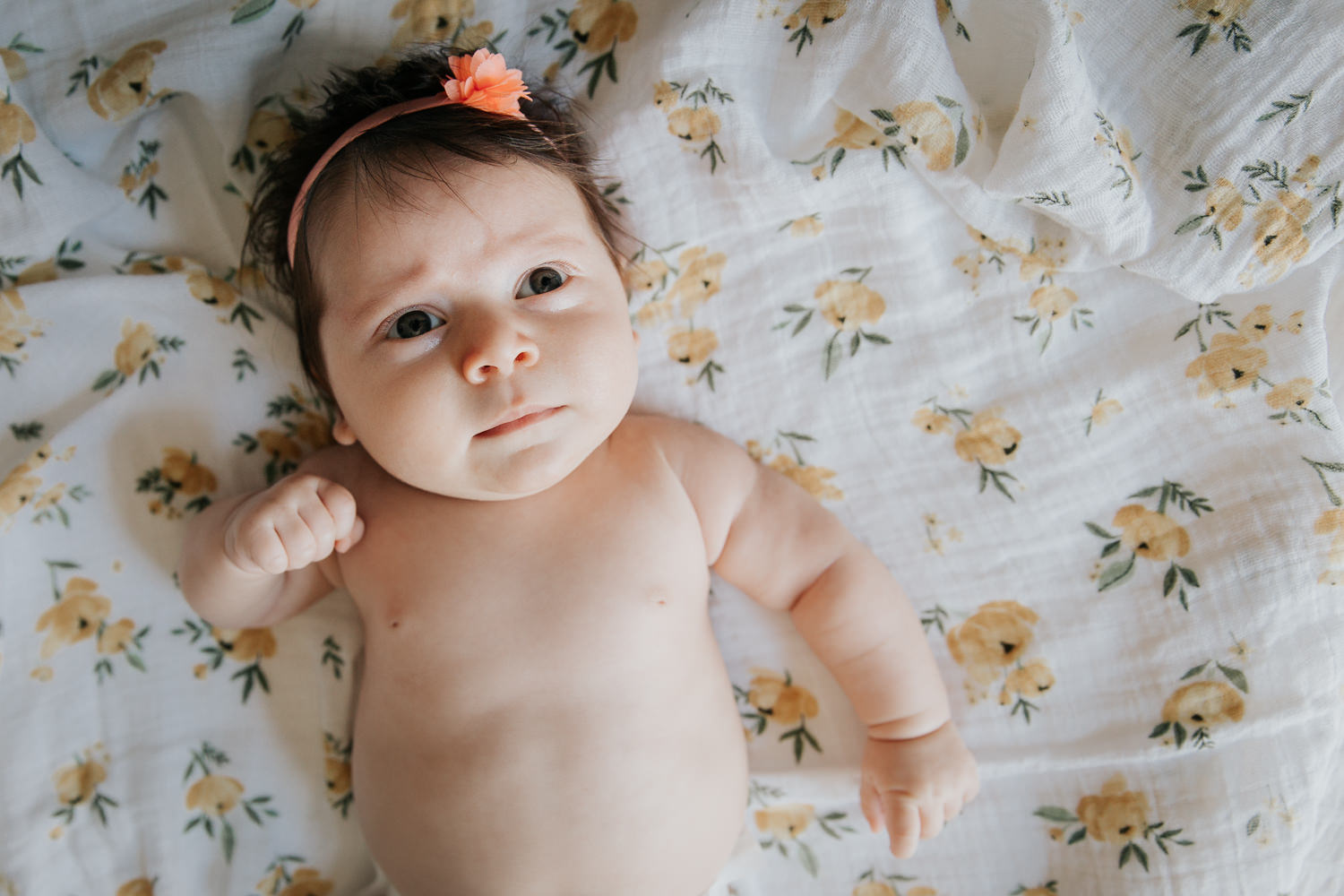 2 month old baby girl in diaper with lots of dark brown hair wearing headband lying on yellow floral blanket, looking perplexed - Markham Lifestyle Photos