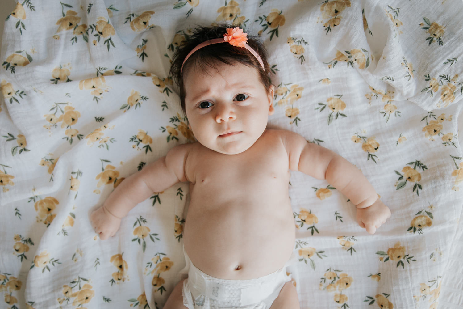 2 month old baby girl in diaper with lots of dark brown hair wearing headband lying on yellow floral blanket, looking perplexed - Newmarket Lifestyle Photography
