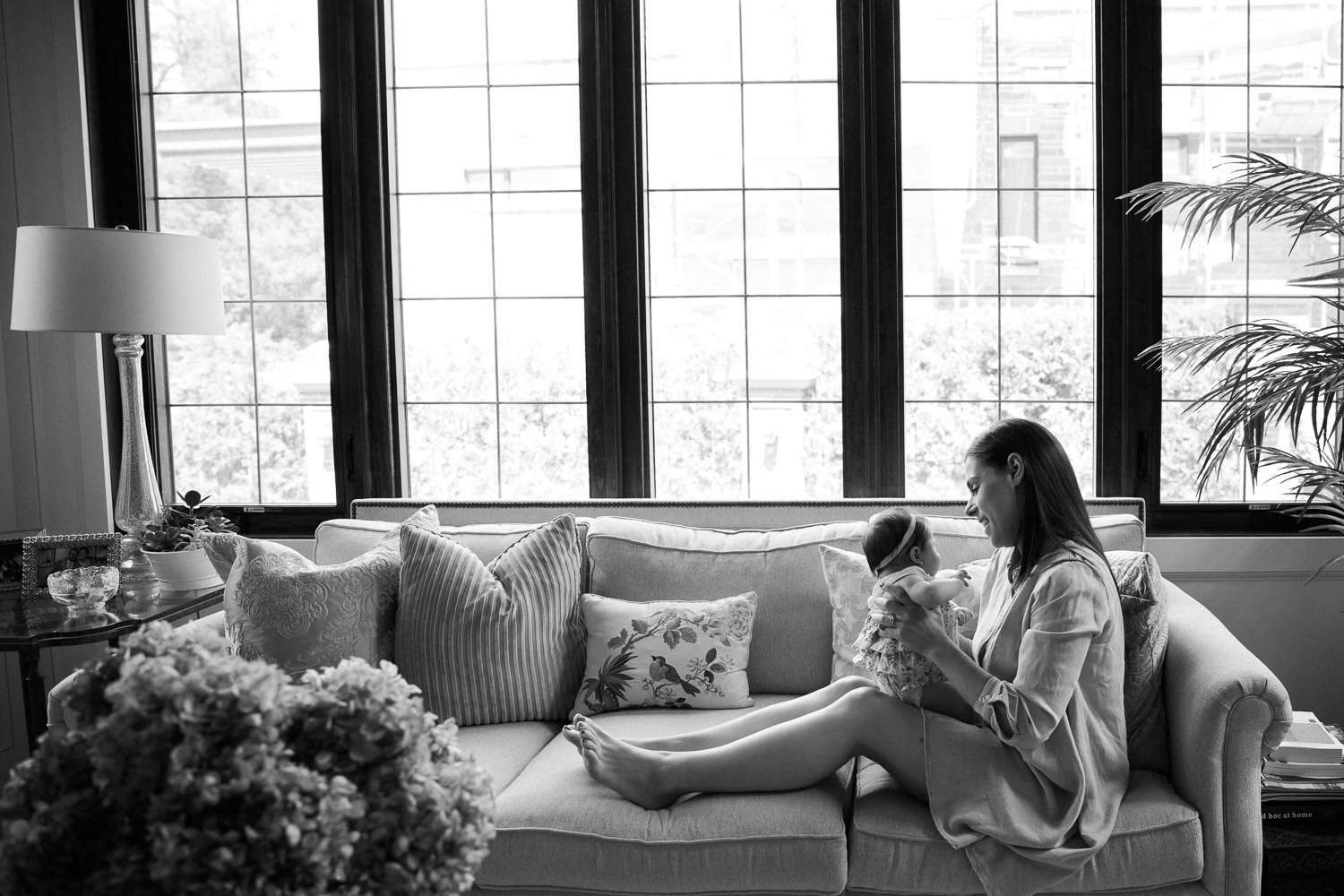 new mom sitting in front of wall of windows with feet up on couch holding and smiling at 2 month old baby girl - Barrie In-Home Photos