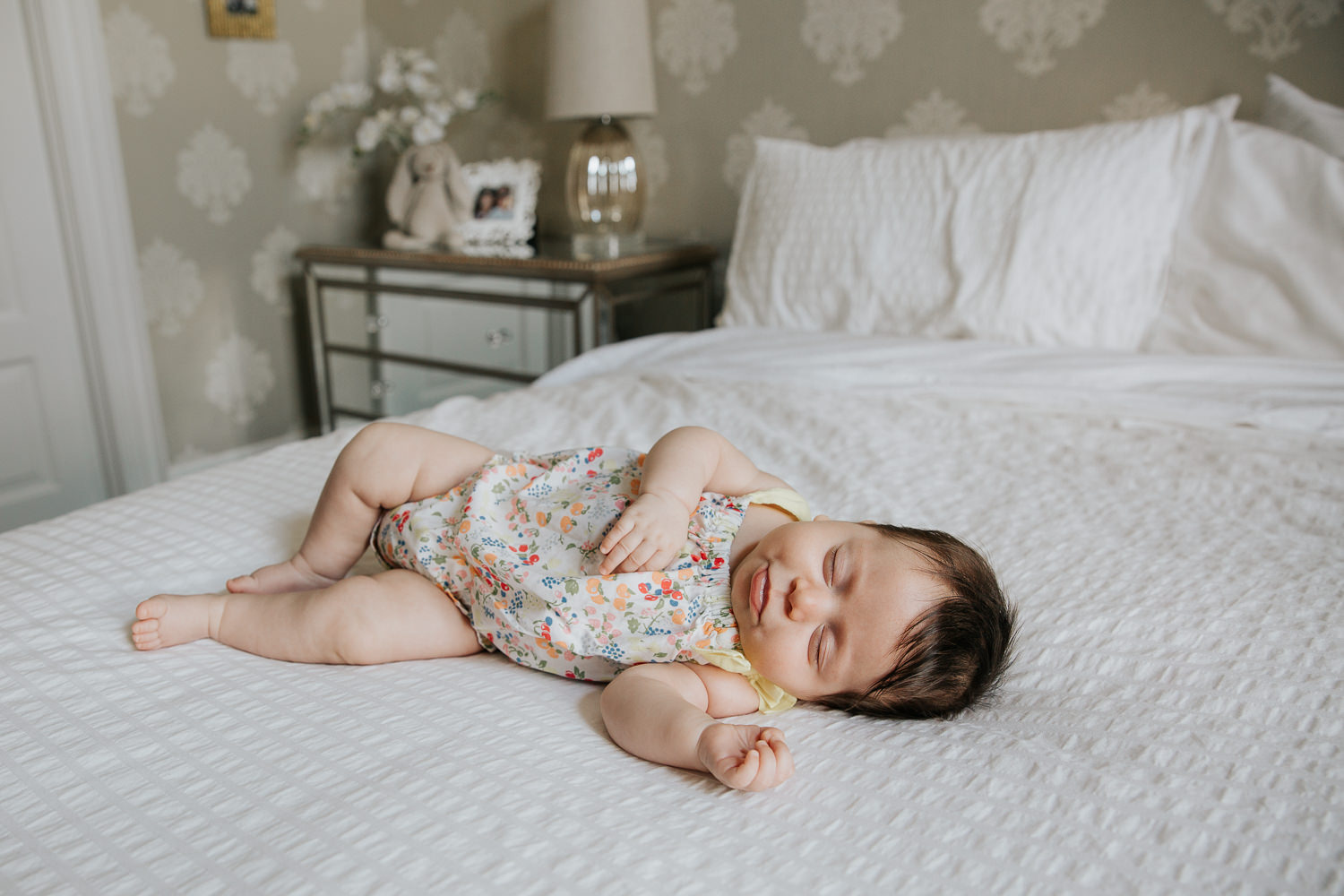 2 month old baby girl with lots of dark hair in floral jumper asleep on bed with arm stretched out - Barrie Lifestyle Photography