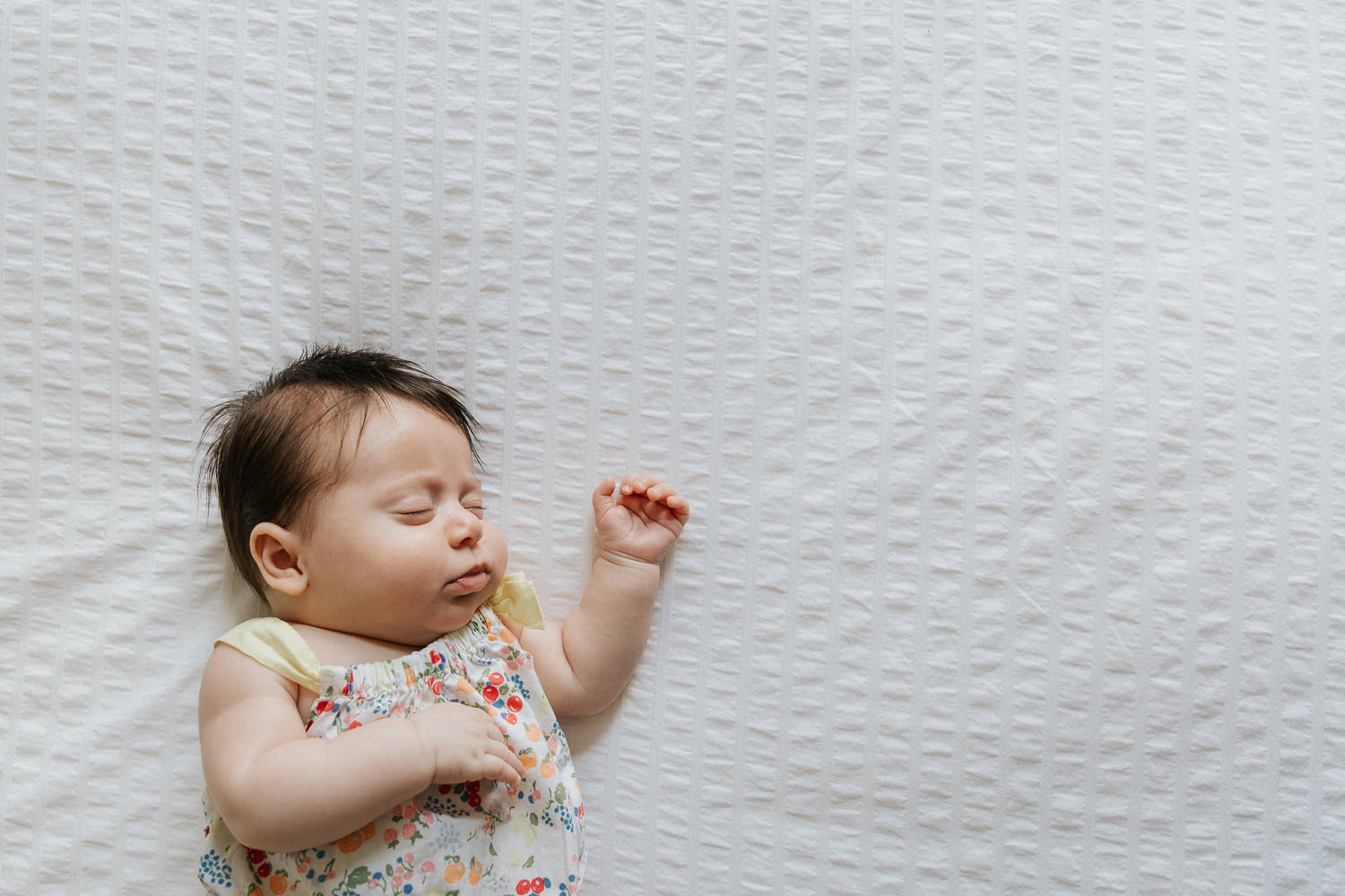 2 month old baby girl with lots of dark hair in floral jumper asleep on bed with arm stretched out - GTA Lifestyle Photography