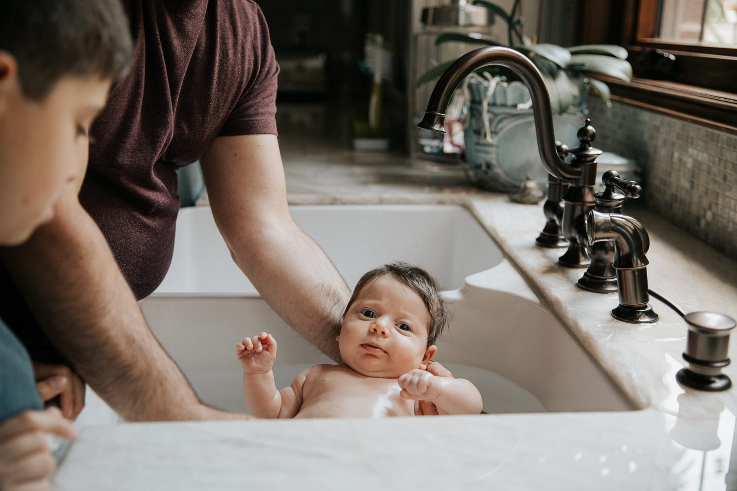 2 month old baby girl with lots of dark brown hair in kitchen sink getting a bath from dad and 9 year old big brother - Newmarket In-Home Photography