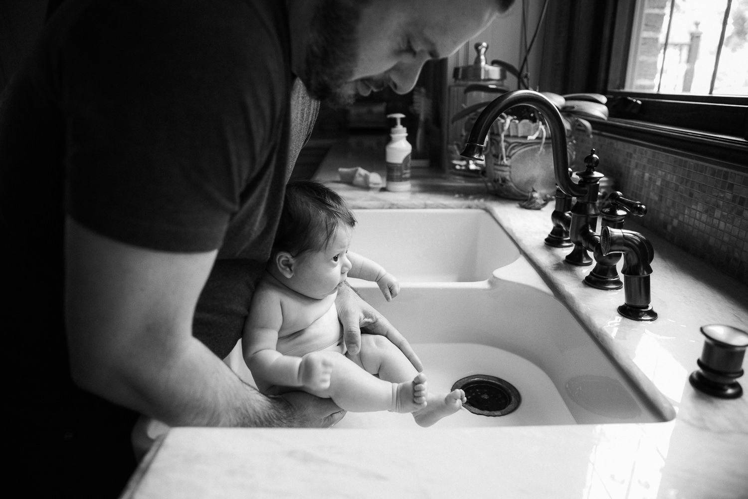 father putting 2 month old baby girl into the kitchen sink for bath time - GTA Lifestyle Photography