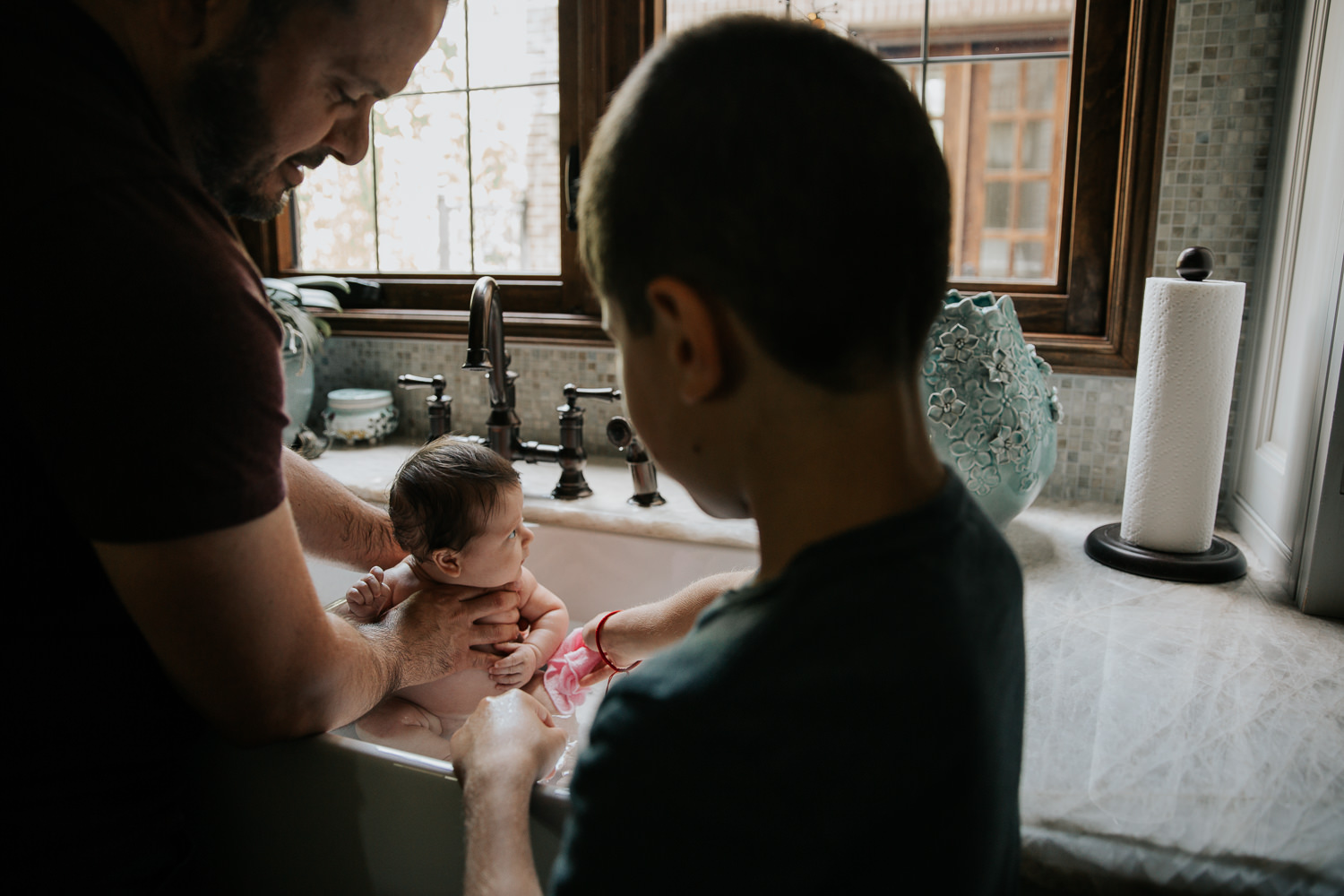 2 month old baby girl with lots of dark brown hair in kitchen sink getting a bath from dad and 9 year old big brother - Markham In-Home Photography