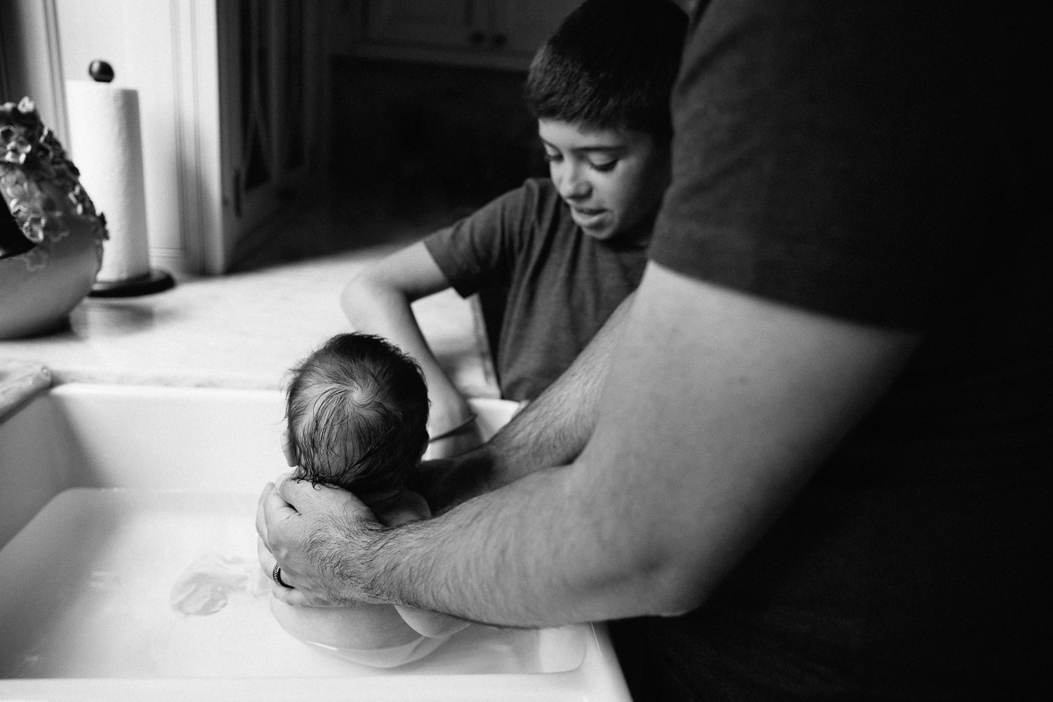 2 month old baby girl with lots of dark brown hair in kitchen sink getting a bath from dad and 9 year old big brother - Barrie In-Home Photography