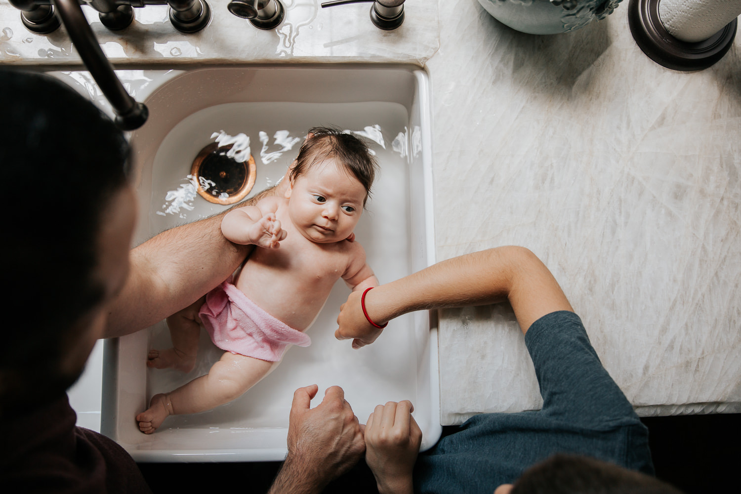 2 month old baby girl with lots of dark brown hair in kitchen sink getting a bath from dad and 9 year old big brother - Newmarket In-Home Photos