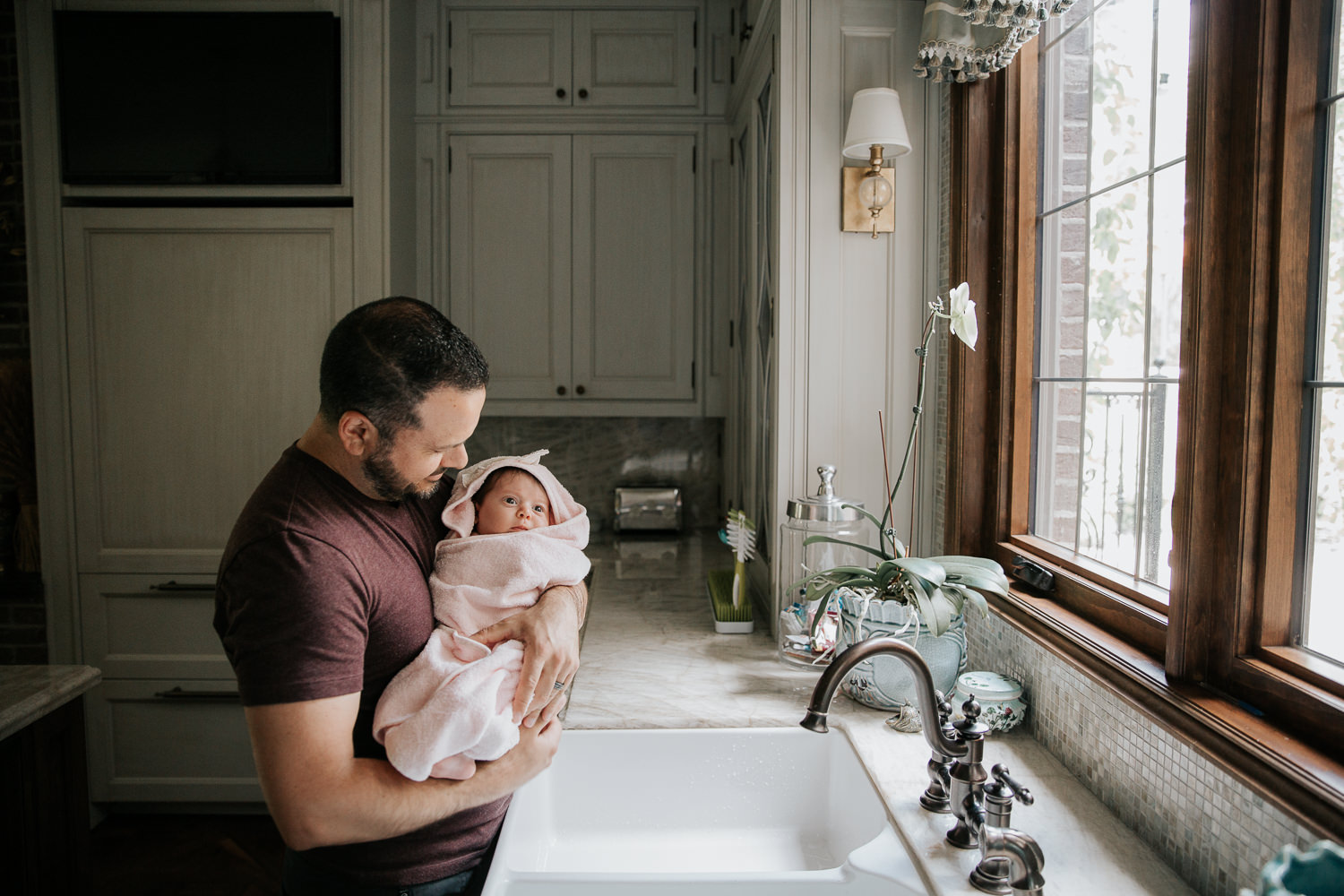 father standing next to kitchen sink, holding 2 month old baby girl wrapped in pink hooded towel after her bath - Markham In-Home Photos