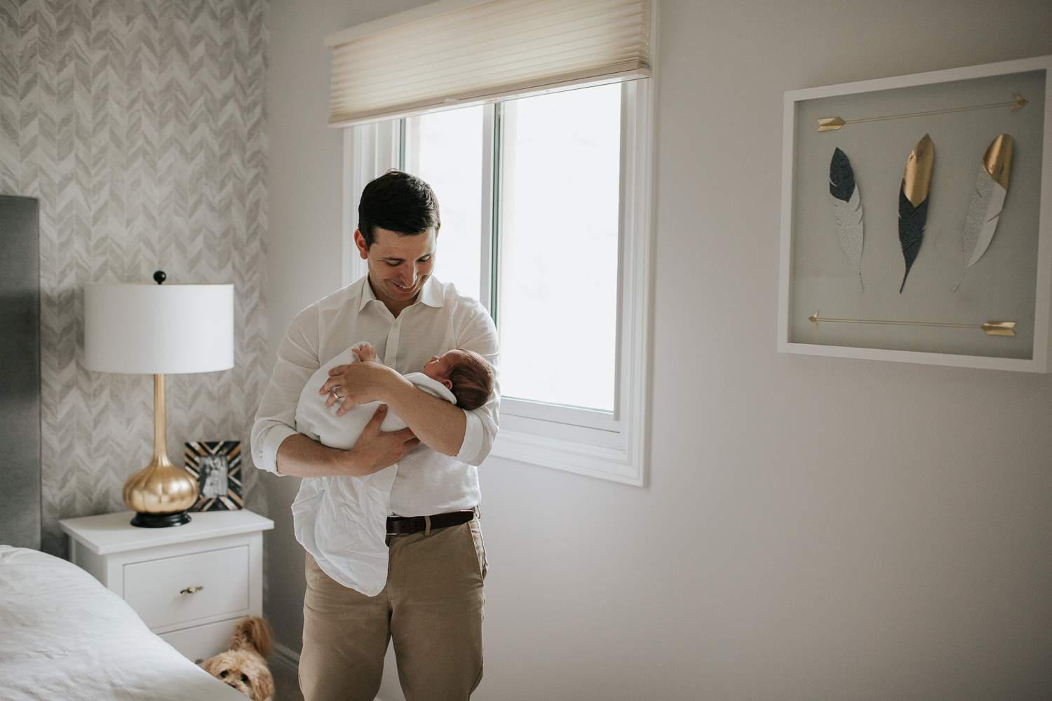 new dad standing next to bed in master bedroom holding 2 week old baby boy in white swaddle, rocking him to sleep - GTA Lifestyle Photography