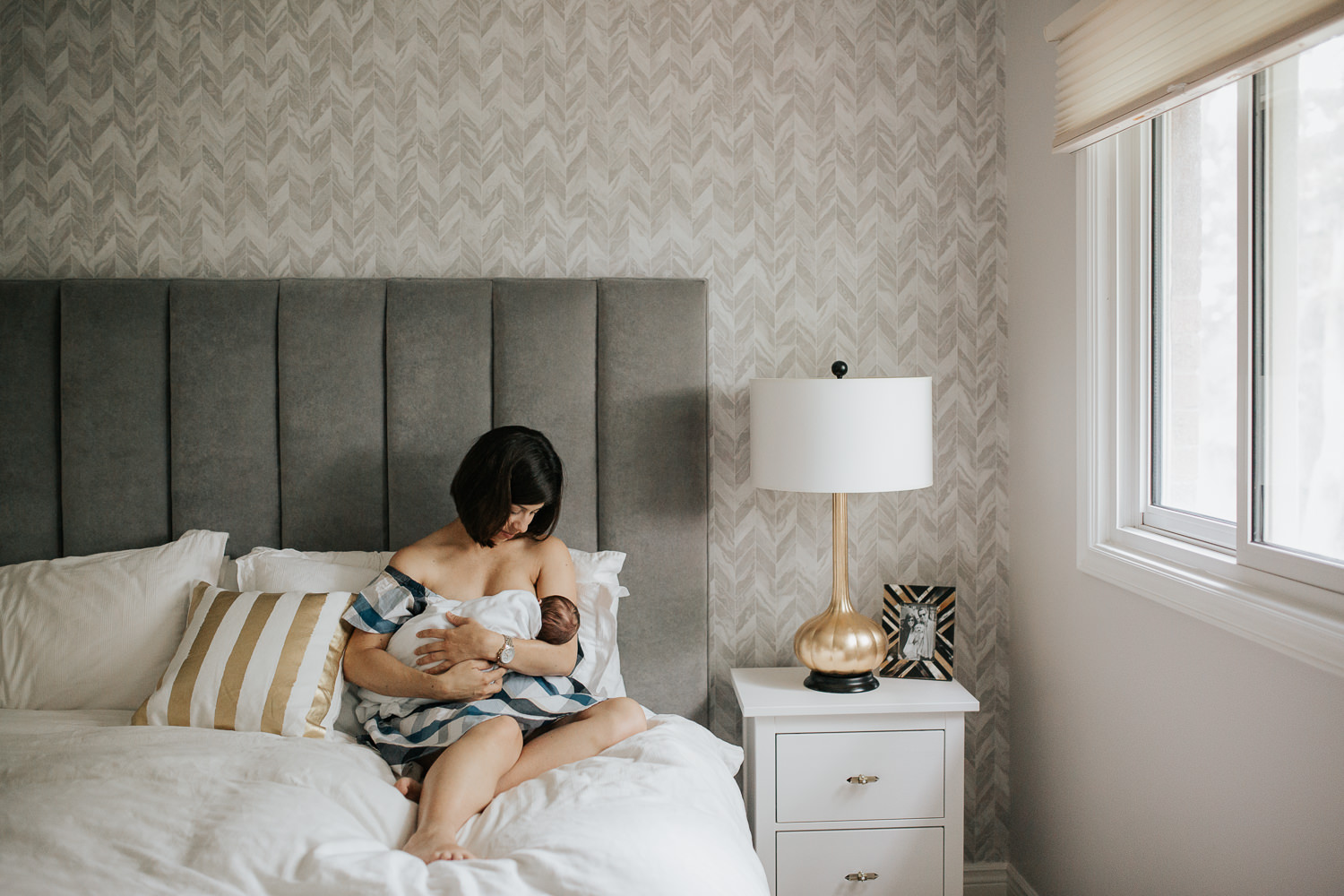 new mom with short brunette hair sitting on master bed nursing 2 week old baby boy in white swaddle - Markham In-Home Photography