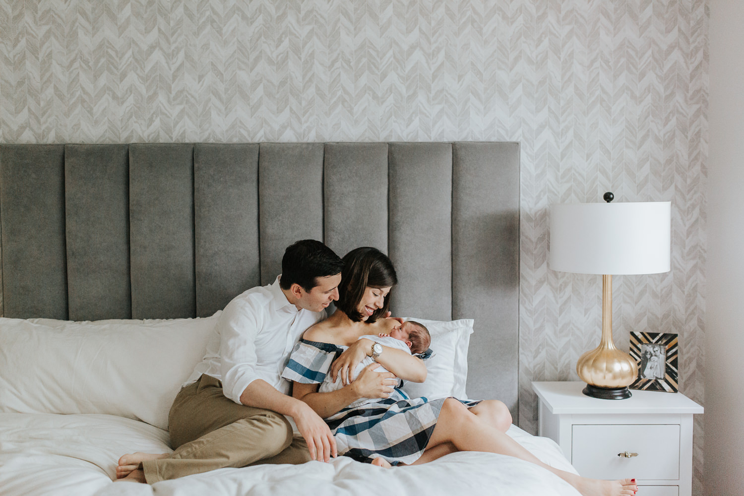 new parents sitting on master bed, mom holding 2 week old baby boy with dad sitting next to them -  Stouffville In-Home Photos
