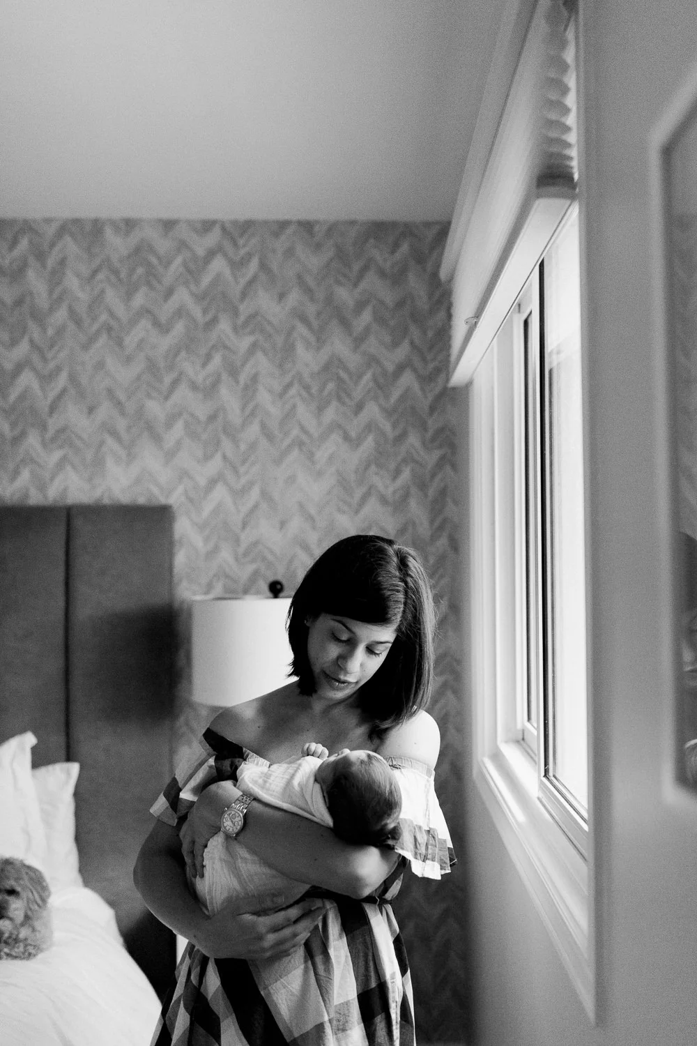 new mother standing next to bedroom window holding and shushing 2 week old baby boy in white swaddle - Markham In-Home Photos