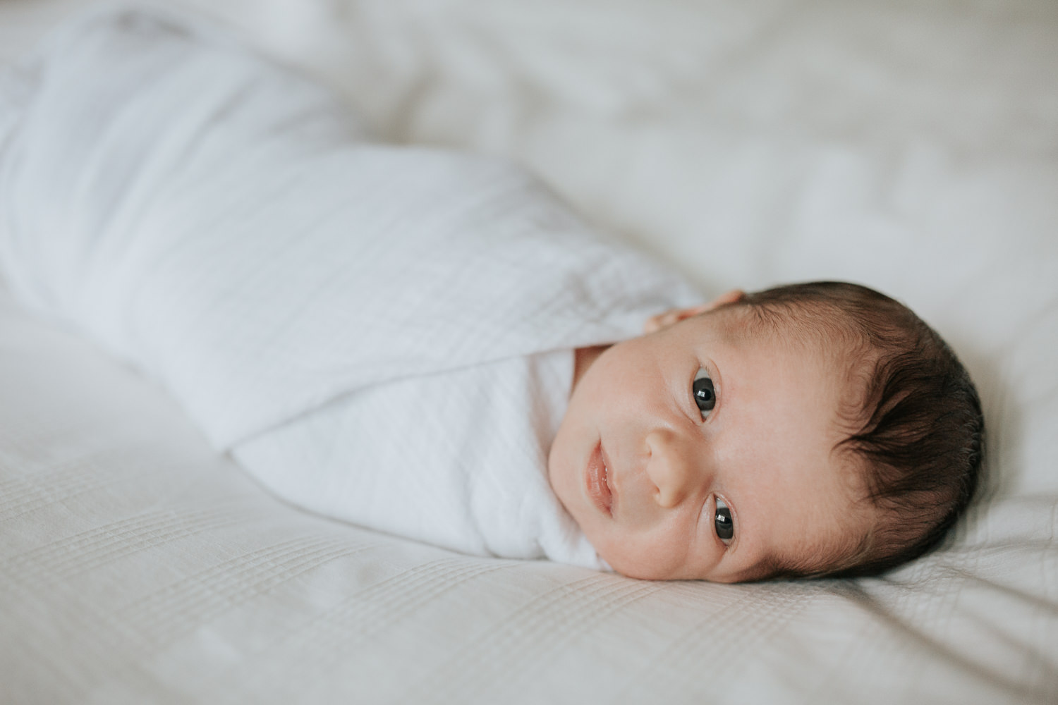 2 week old baby boy with dark hair in white swaddle lying on bed awake looking at camera - Stouffville Lifestyle Photography