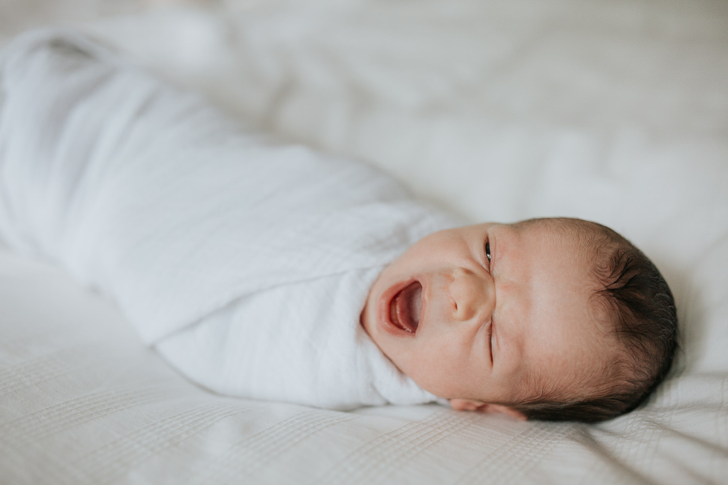 2 week old baby boy with dark hair in white swaddle lying on bed yawning - Markham Lifestyle Photography