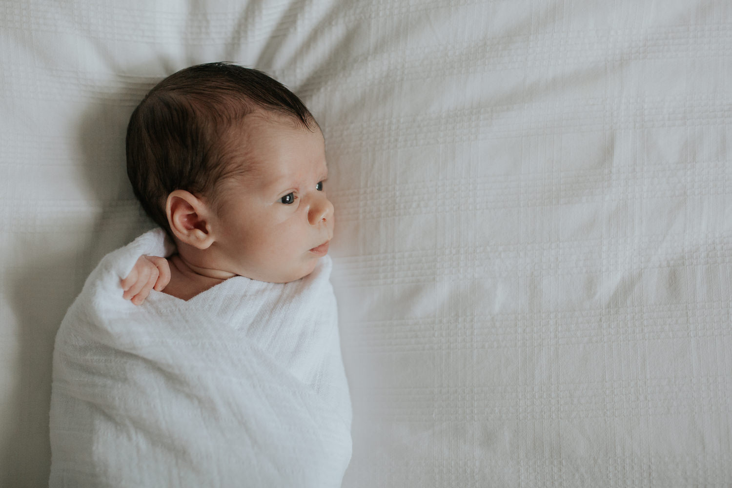 2 week old baby boy with dark hair in white swaddle lying on bed awake looking to the side - Barrie Lifestyle Photography