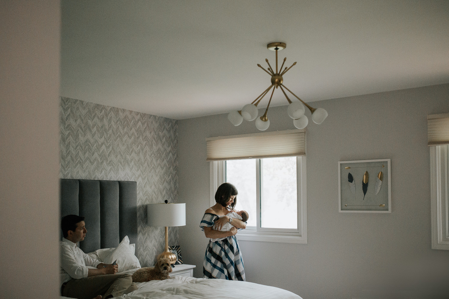 new mother standing next to bedroom window holding 2 week old baby boy in white swaddle, dad and dog sitting on bed - GTA In-Home Photos