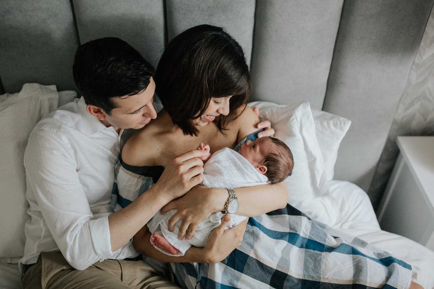 new parents sitting on master bed, mom holding 2 week old baby boy in white swaddle and dad holding his hand - Stouffville Lifestyle Photos