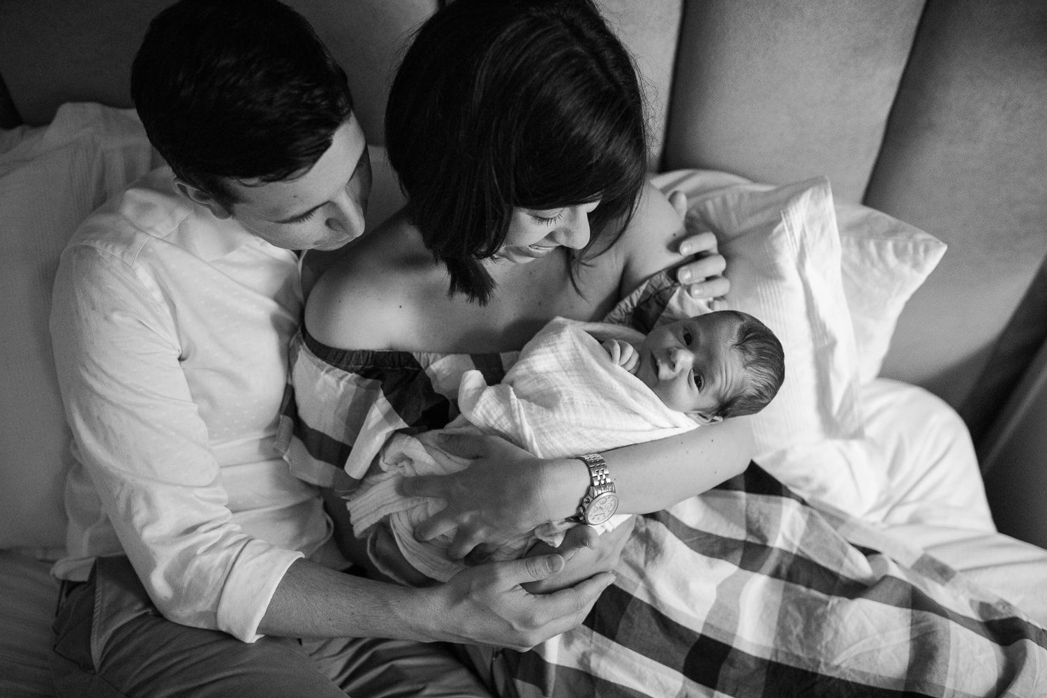new parents sitting on master bed, mom holding 2 week old baby boy in white swaddle and dad embracing them - Markham Lifestyle Photos