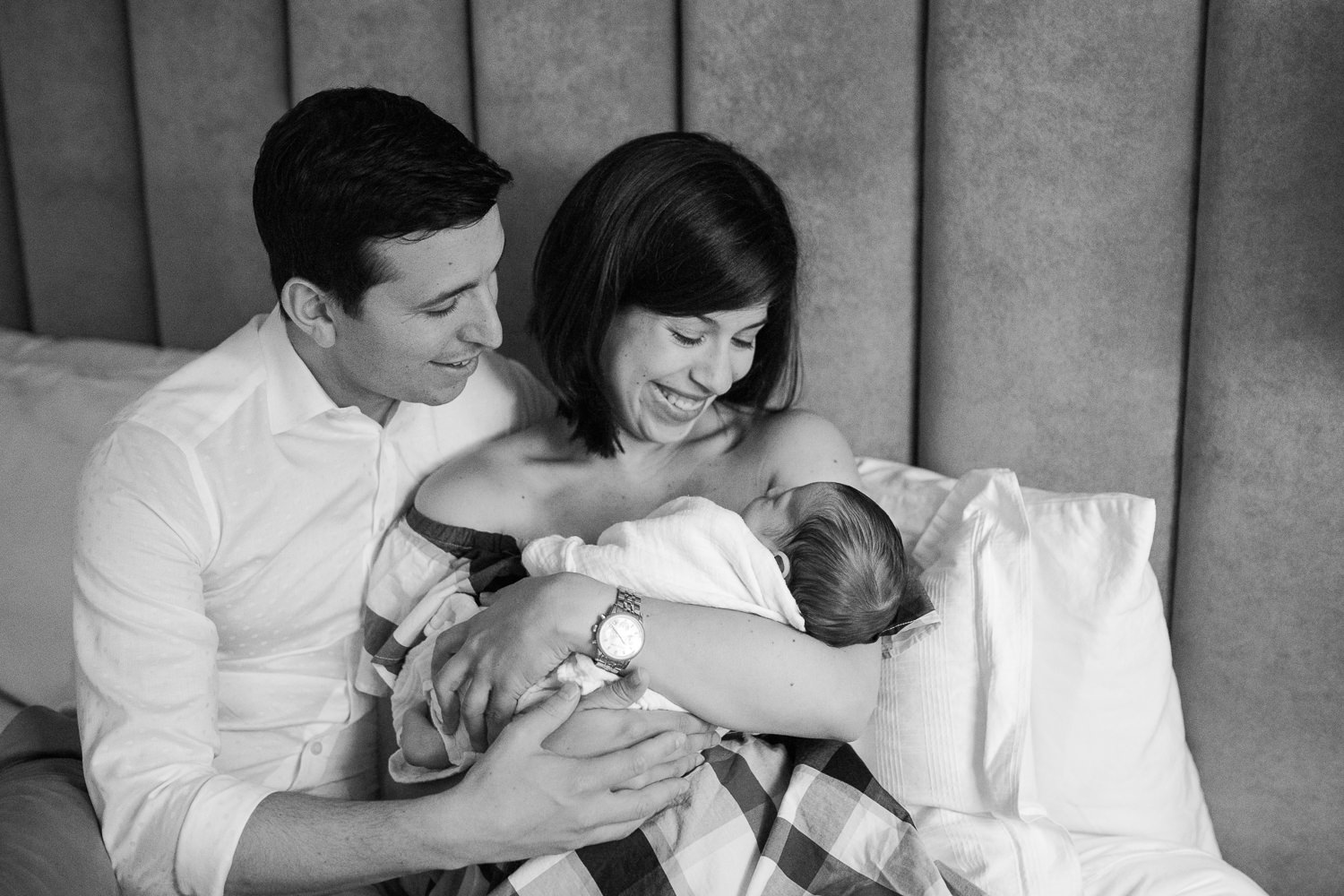 new parents smiling sitting on master bed, mom holding 2 week old baby boy in white swaddle and dad embracing them - Barrie Lifestyle Photos