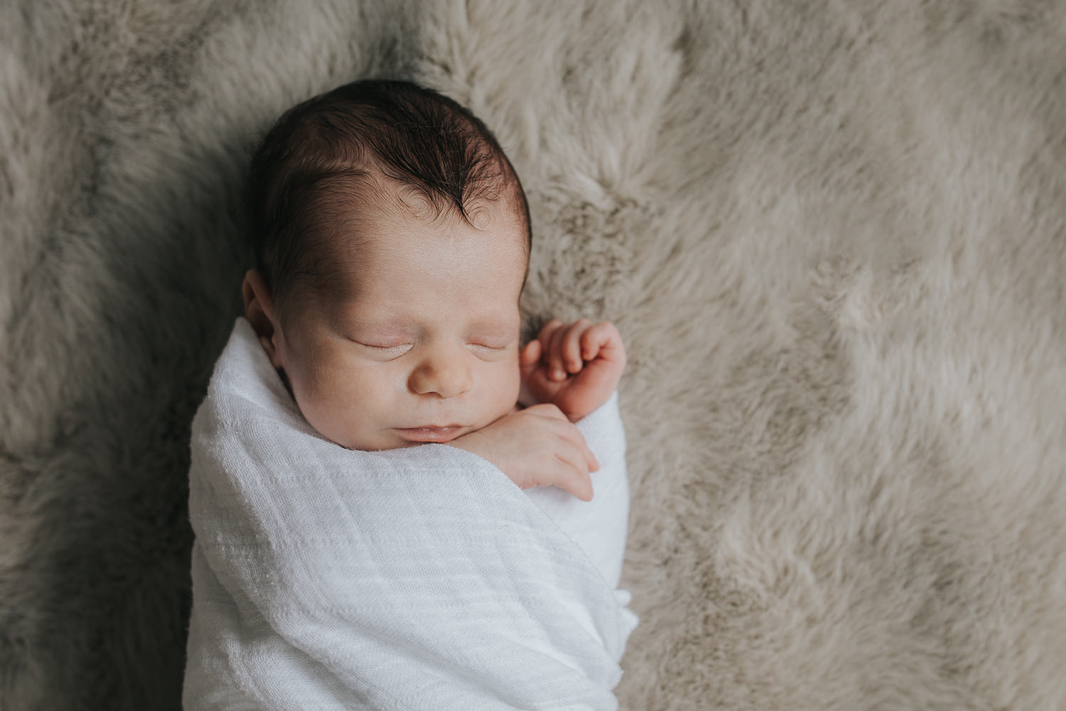 2 week old baby boy with dark hair lying on fur blanket in white swaddle, sleeping with hands next to face - Stouffville Lifestyle Photography